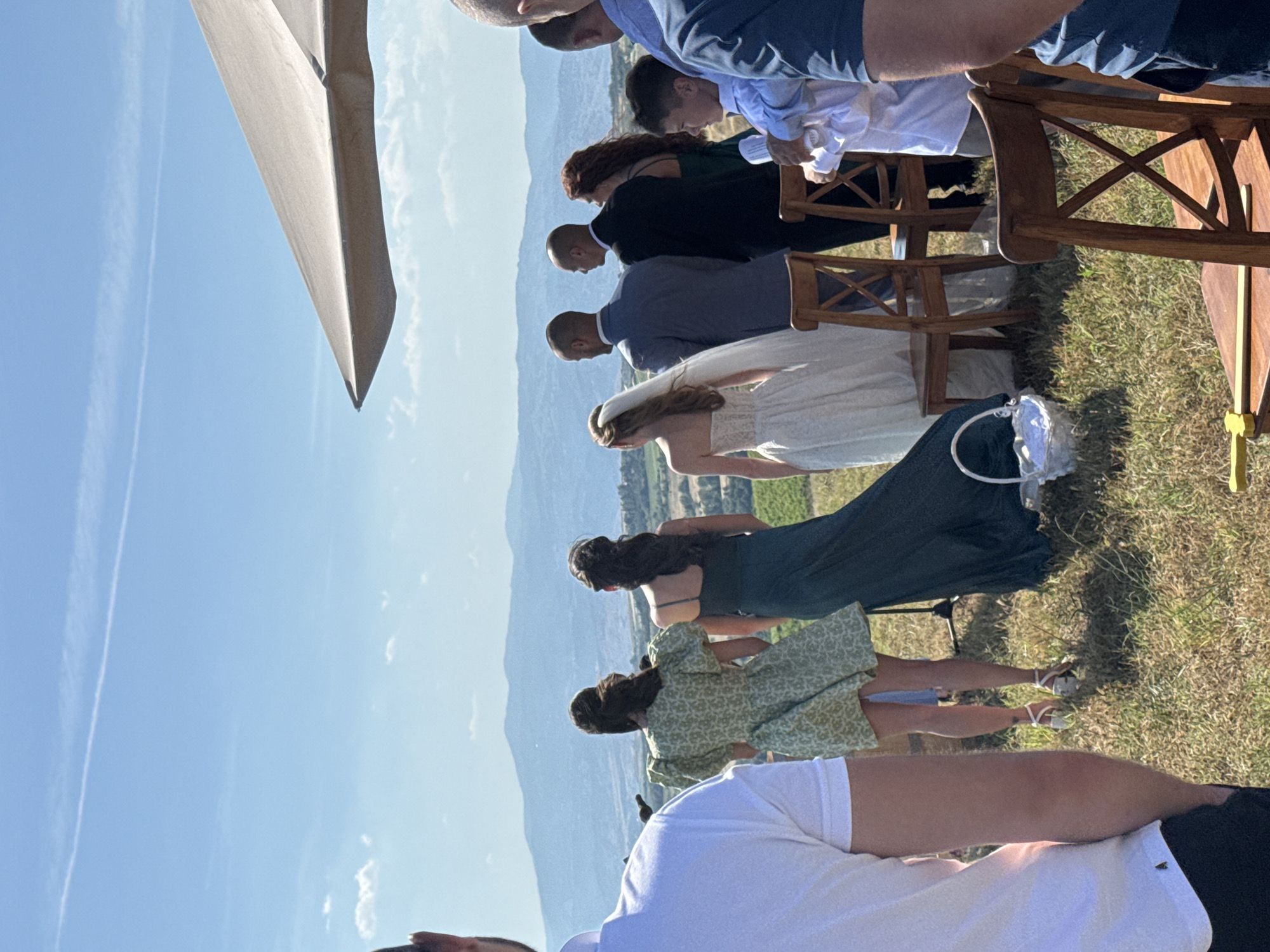 An outdoor gathering, possibly a wedding, is observed from behind a group of people, under a bright blue sky in the city of Harsovo, Bulgaria. The scene unfolds on a sunlit, grassy hill overlooking a vast landscape of hazy blue mountains and green fields in the distance.
In the immediate foreground on the left, the back and right arm of a man in a white polo shirt and dark pants are visible. On the right, a large, light-colored umbrella extends into the frame, providing partial shade. Two empty wooden chairs with X-shaped backrests are positioned on the dry, yellowish-green ground. A yellow stick-like object rests on the seat of the chair closer to the viewer.
A line of at least seven individuals stands in the mid-ground, all facing away from the camera towards the panoramic view. From left to right, a woman in a short green patterned dress with puff sleeves and light-colored high heels stands next to a woman in a long, dark green dress. Following them is a bride in a white strapless wedding gown with a veil, holding a white basket on the ground beside her. Next are two men in suits, one in a light blue jacket and another in a dark jacket. A woman with long red hair in a dark dress is partially visible behind the second man. To their right, a young boy in a white long-sleeved shirt looks towards the left, and a man in a blue shirt is partially visible on the far right. The ground is dry and uneven, with patches of dry grass and dirt. The background features rolling, hazy blue mountains under a clear sky with a single faint contrail and a few scattered white clouds.