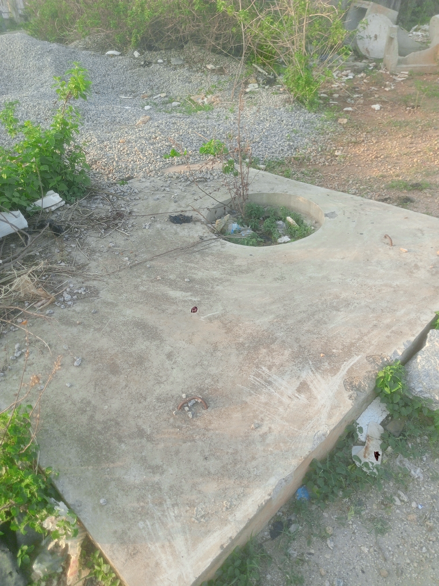 The image shows a concrete structure with a circular opening in the center, located in Gwarinpa, Nigeria. The structure appears weathered and stained. The circular opening contains debris, including trash and plant growth. Vegetation grows around the edges of the concrete structure and in the surrounding area, which includes gravel and dirt. Remnants of construction or demolition are visible in the background.