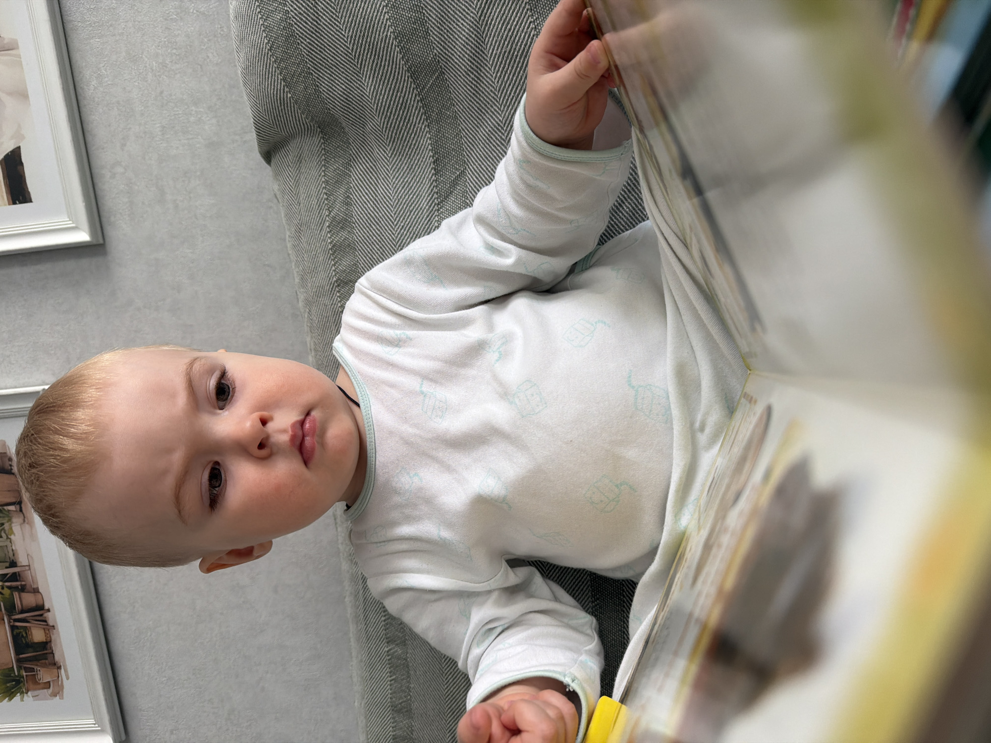 A baby wearing a white long-sleeved shirt with light blue patterns is lying on a light gray sofa or cushion with a herringbone pattern. The baby is holding a book open in front of them with their right hand, partially obscuring the view of the room behind. A framed picture hangs on the wall to the left of the sofa. The picture's content is partially visible, showing what appears to be a building or landscape. The photo's details suggest an interior setting, possibly a private residence. The location, Votkinsk, Russia, is not discernible from visual details in the image.