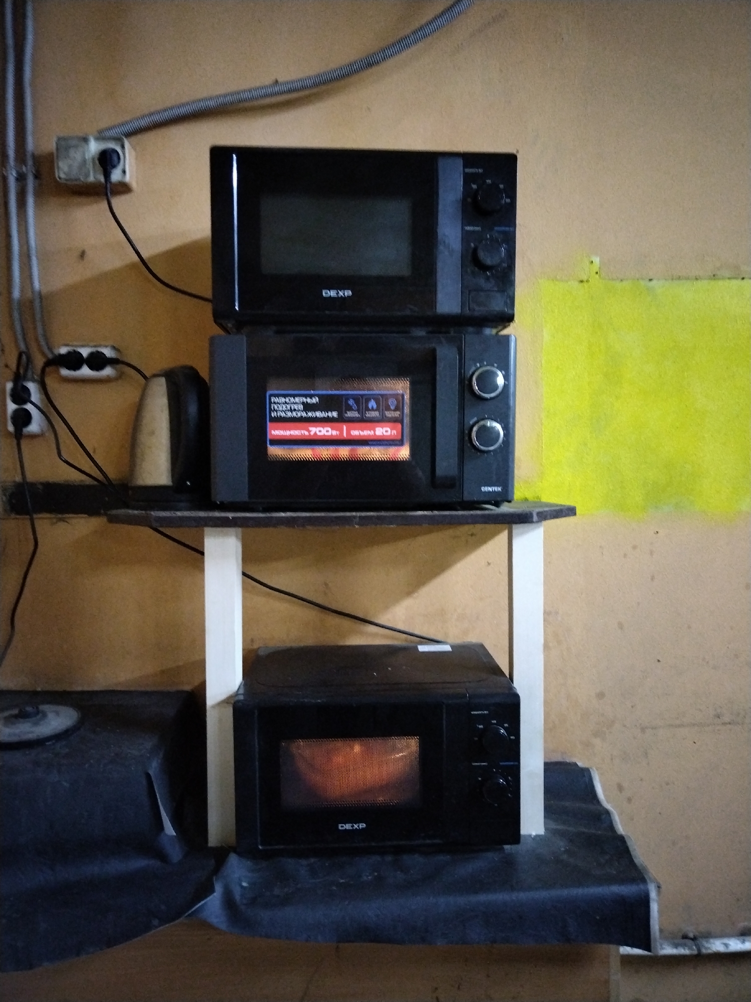 Three black microwave ovens are stacked vertically in a room with tan walls. The top microwave is open on the left side. The middle microwave has a screen displaying text and icons, and two silver knobs on the right. The bottom microwave has its door closed and the interior is lit. The microwaves are on a dark shelf supported by white vertical poles. In the background, a yellow painted area is visible on the wall. Electrical outlets and wiring are present on the left side of the image. The scene appears to be a storage area or workshop.