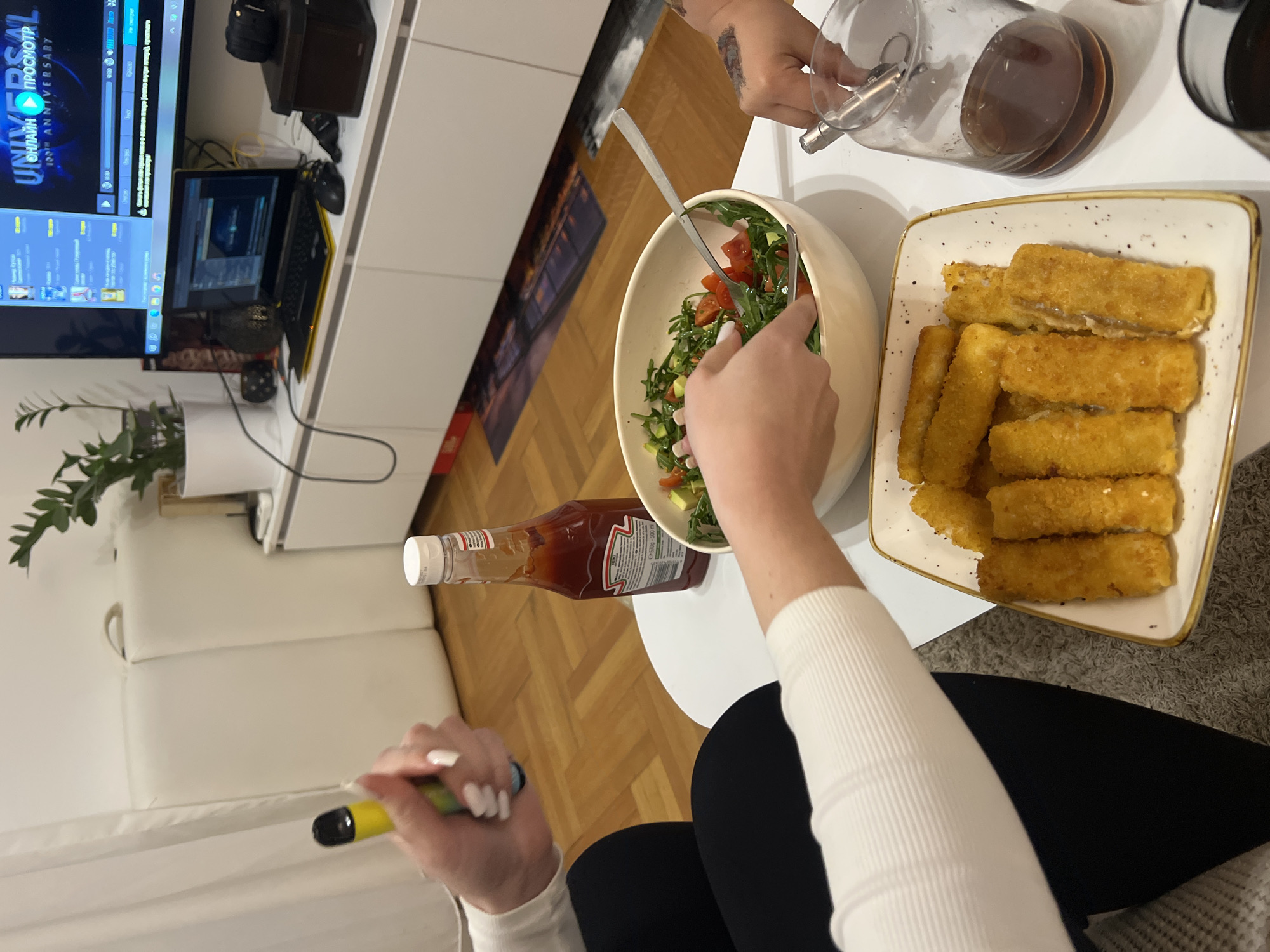 The scene is indoors, featuring a person seated at a round white table with a salad, fried sticks, a ketchup bottle, and a carafe of liquid. The person is wearing black pants and a white long-sleeved shirt, holding a vape pen in their left hand and using salad servers to mix the salad. Another hand is visible pouring liquid from a carafe. The table is set in front of a white couch. In the background, there is a TV displaying content, a laptop, and various decorative items on a white shelf. The floor is made of light brown parquet. The location appears to be a private residence, not specifically identifiable as being in Budva, Montenegro without further evidence.