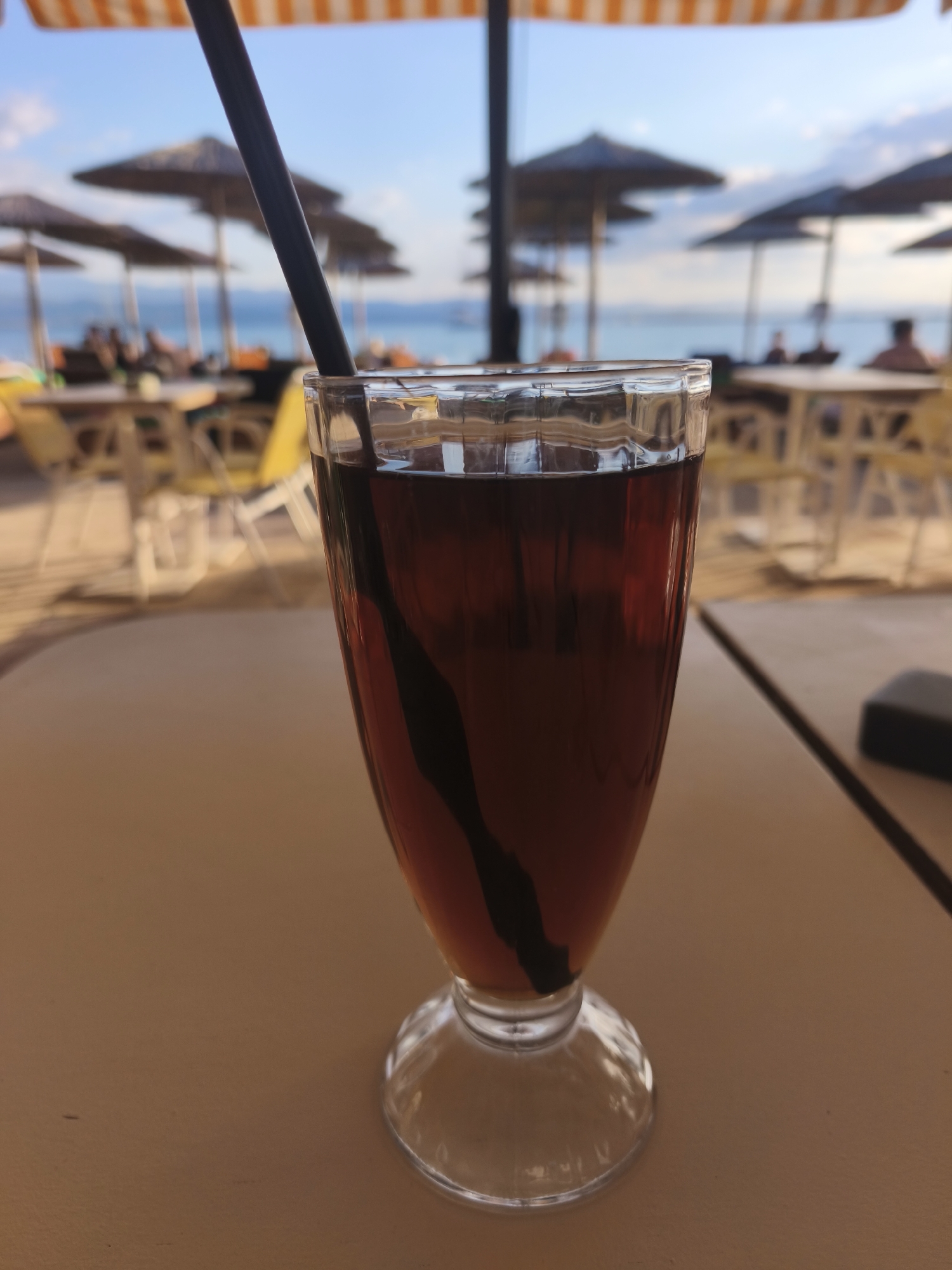 A close-up, medium shot shows a glass filled with a dark liquid and a black straw, resting on a light brown surface. The glass has a pedestal base. In the background, out of focus, is a beach scene with numerous umbrella-covered lounge chairs, tables, and people relaxing. The sky is a pale blue with some clouds, and the sea is visible in the distance. The setting appears to be an outdoor cafe or restaurant at a beach resort in Eretria Municipal Unit, Greece.