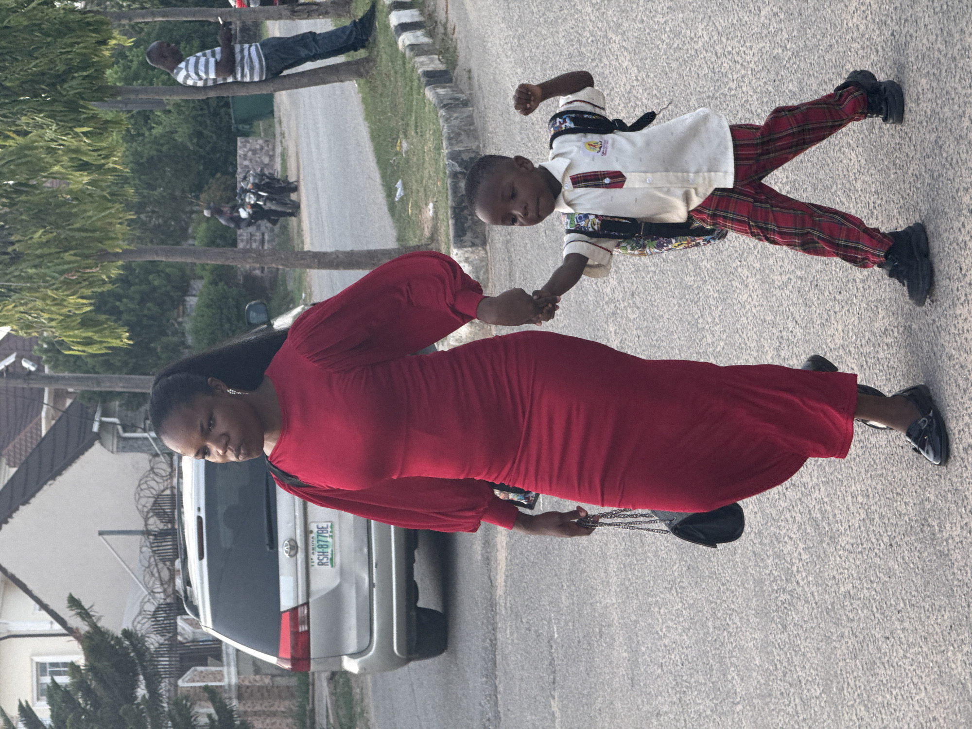 The image captures a street scene in Abuja, Nigeria, during what appears to be late afternoon or early evening. The primary subjects are a woman and a young boy walking hand-in-hand on a paved road, which appears rotated sideways in the frame.
The woman, on the left side of the image, is dark-skinned with dark hair pulled back. She wears a long-sleeved, floor-length red dress and black flat shoes. In her left hand, she carries a black purse with a chain strap. Her expression is neutral as she looks towards the camera.
The young boy, walking beside her and holding her hand, is also dark-skinned with short dark hair. He is dressed in a light-colored collared shirt with plaid (red and black) detailing and a tie or strap across the chest. He wears matching red plaid trousers and black shoes, possibly sneakers. A patterned backpack or bag is visible on his back. His gaze is directed forward.
In the background, to the right, a man with dark skin is seen standing under a tree, wearing a striped shirt and blue jeans, looking down at a mobile phone. Further in the background, several motorcycles are parked.
A silver Toyota vehicle, possibly a minivan or SUV, is parked along the side of the road, displaying a Nigerian license plate that reads "LAG ABUJA RSA-877BE". Houses with light-colored walls and dark roofs are visible behind the parked car, with barbed wire fencing on a wall. The street is lined with green trees. The overall mood is calm and depicts a casual, everyday activity.