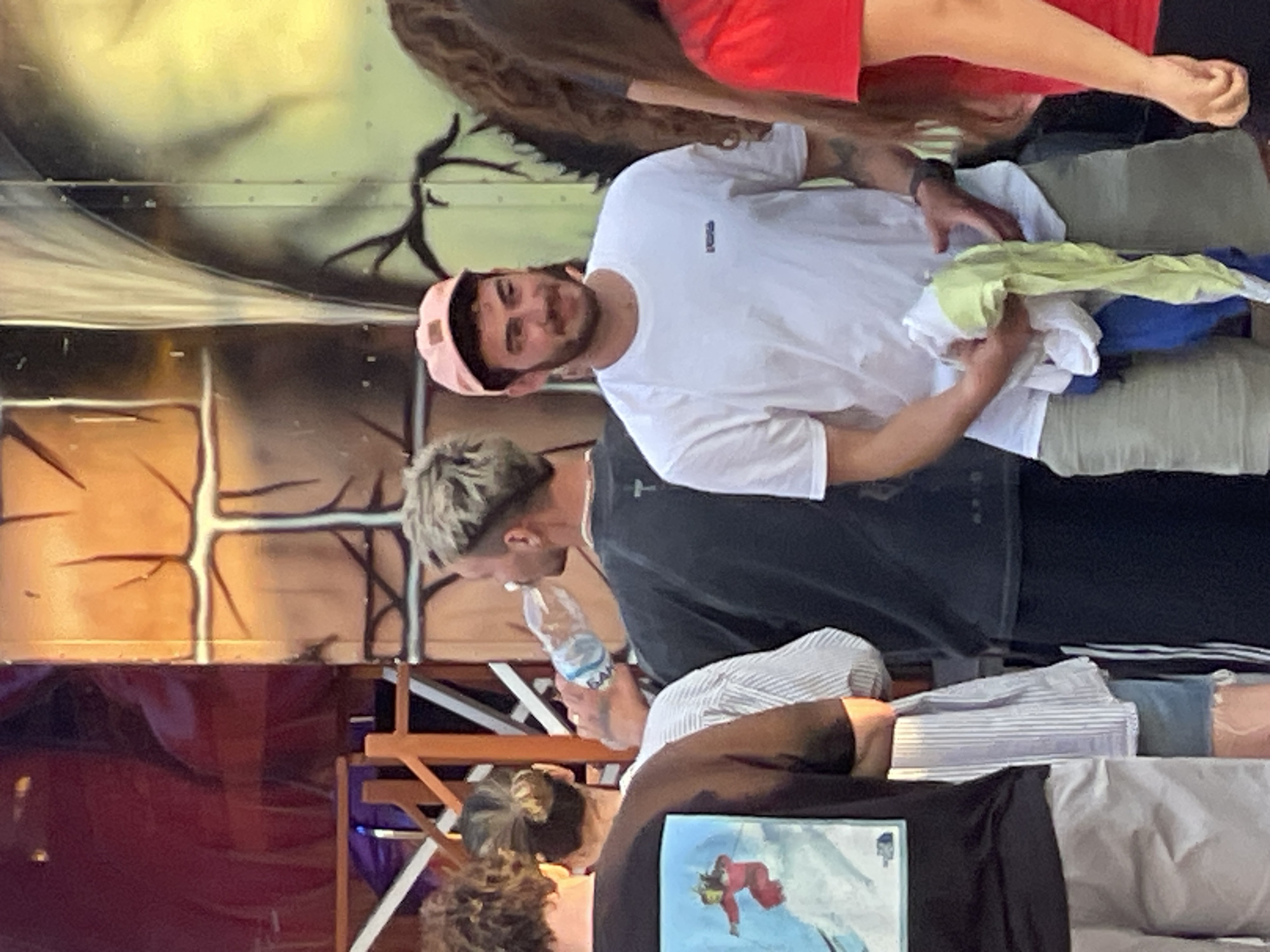 At an outdoor event, possibly an amusement park or festival, in Varna, Bulgaria, a group of people are gathered. The focal point is a young man wearing a pink baseball cap and a white t-shirt, holding a bundle of colorful cloths. To his left, another man with blonde hair is drinking from a water bottle. Behind them, parts of a structure with a rough, textured facade painted with cracks and dark figures are visible. The overall lighting suggests it is daytime, likely late afternoon or early evening, with a warm, diffused light.