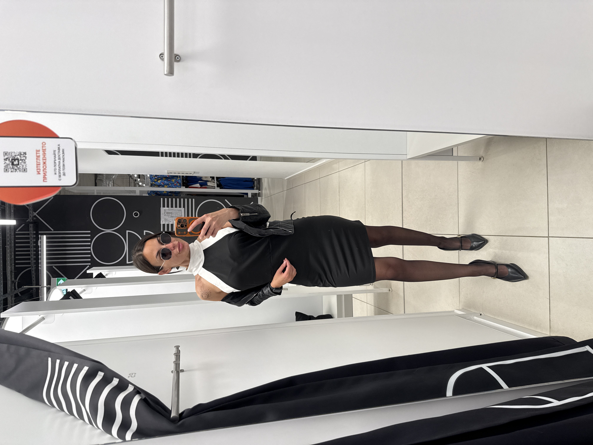 A person wearing sunglasses, a black dress, black tights, and black shoes is taking a selfie in what appears to be a dressing room. The person is holding a phone and looking at the reflection in the mirror. A black leather jacket is draped over the outfit. A QR code is visible in the upper left of the frame. Clothes are hanging in the foreground. The background includes store decor with black and white patterns. While the prompt specifies Pleven, Bulgaria, there is nothing in the image to verify the location is Pleven or Bulgaria.