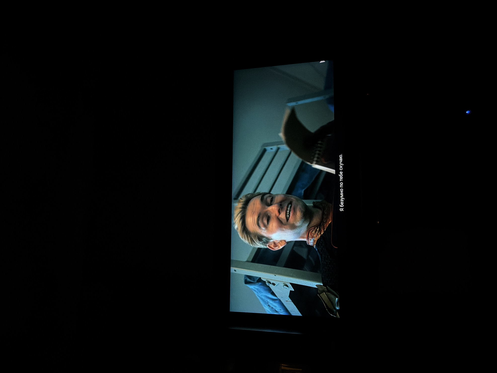 The image shows a close-up shot of a man with blond hair and a slightly deranged expression on his face, presumably taken from a television or computer screen. He is wearing a dark outfit with some decorative elements around the collar. In the background, there is a partially visible structure that appears to be a set of metal shelves or a ladder. Additionally, there is a caption in Russian on the right side of the screen that says "Я безумно по тебе скучаю". The surrounding environment is dark, with the screen being the main source of light. There is no visible information to indicate that the location is in Moscow, Russia.