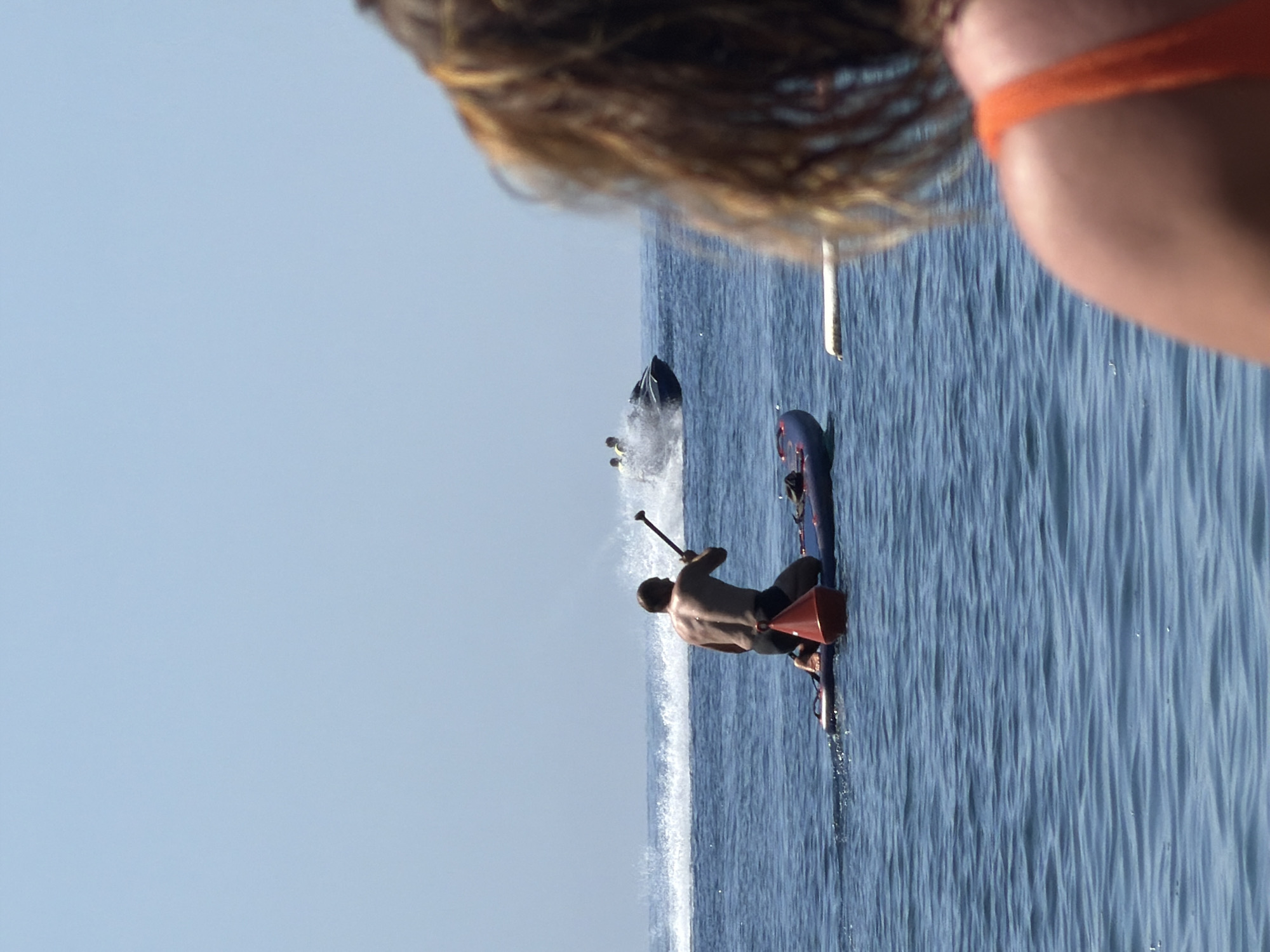 The image displays a daytime scene on the sea in Budva, Montenegro, under a clear, light blue sky. In the extreme foreground, the blurred back of a person's head with light brown hair and a bare right shoulder wearing an orange strap is visible, positioned on the right side of the frame. In the midground, a shirtless male, wearing dark swim shorts, kneels on a blue and purple stand-up paddleboard, holding a paddle. A red conical buoy is attached to the front section of his paddleboard. In the background, a jet ski with two occupants is observed moving rapidly from left to right, creating a significant white wake and spray on the water's surface. The sea exhibits a moderately calm texture with discernible ripples.