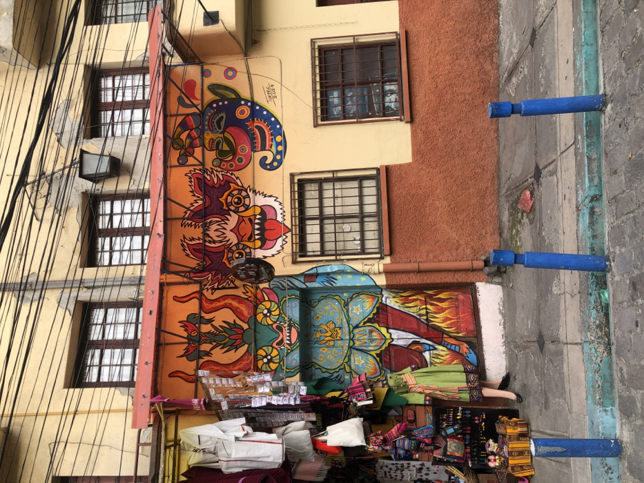 A vibrant mural decorates the side of a building, featuring colorful, stylized creatures. Below the mural, a street vendor displays various textiles and handicrafts, filling the sidewalk with merchandise. The building has multiple windows with dark grilles. Utility wires crisscross the upper left corner. Blue barriers line the edge of the sidewalk. The adjacent building is orange.