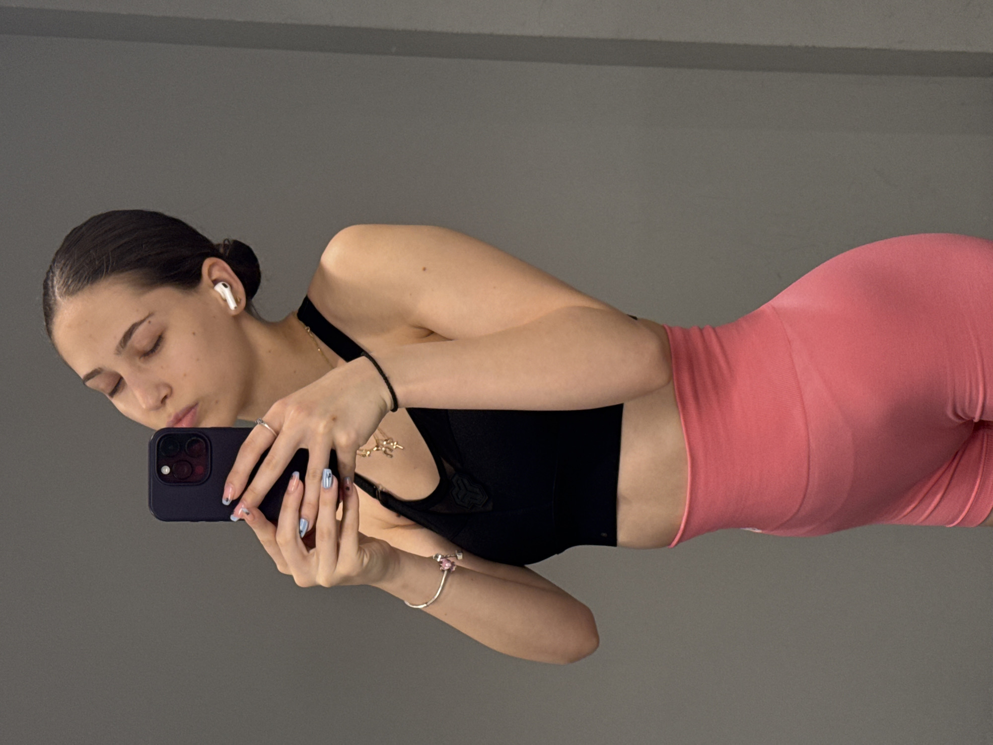 A young woman is posing for a selfie. She is wearing a black sports bra and pink workout shorts. She has dark hair pulled back in a bun and is wearing an earbud. She is holding a dark-colored smartphone with painted nails. She is also wearing a necklace and bracelets. The background is a gray wall. There is nothing to indicate that the location is Sofia, Bulgaria.