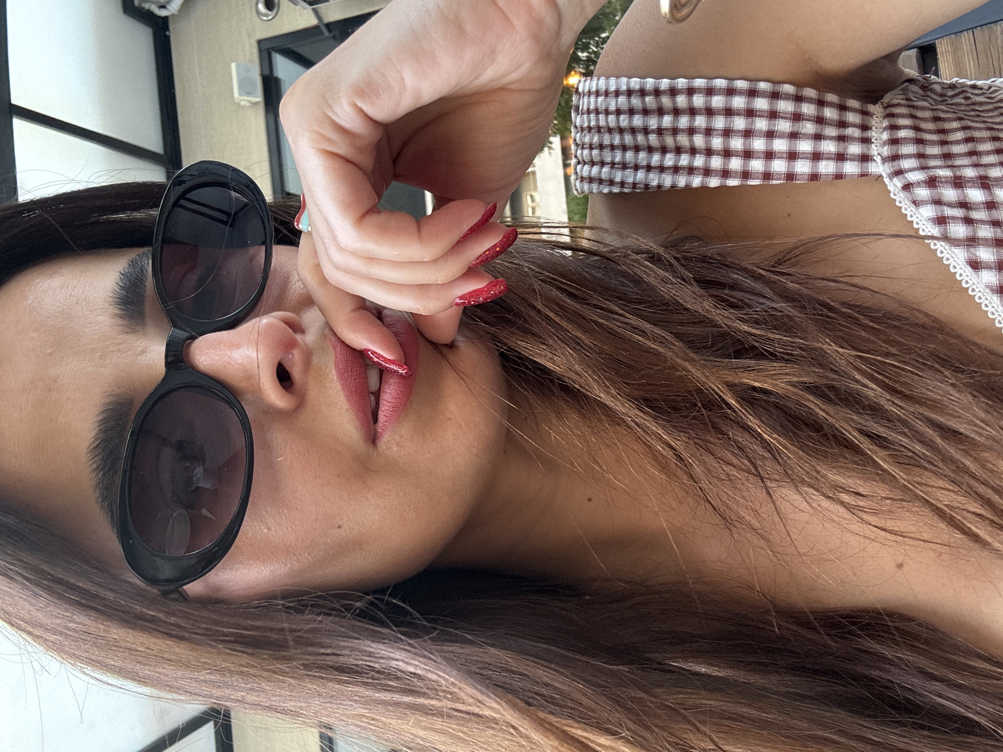 A medium shot of a woman's face and upper body is captured from a slightly low angle. She is wearing large, black, oval-shaped sunglasses. Her right hand is brought up to her mouth, with her fingers resting on her lips. Her fingernails are painted a vibrant red with glitter. She has long, brown hair that falls around her shoulders and face. Her lips are painted a matte pinkish-red. She is wearing a gingham-patterned top with white trim. The background appears to be outdoors, possibly a patio or deck, with a blurred background of structures and foliage. The lighting suggests it is daytime.