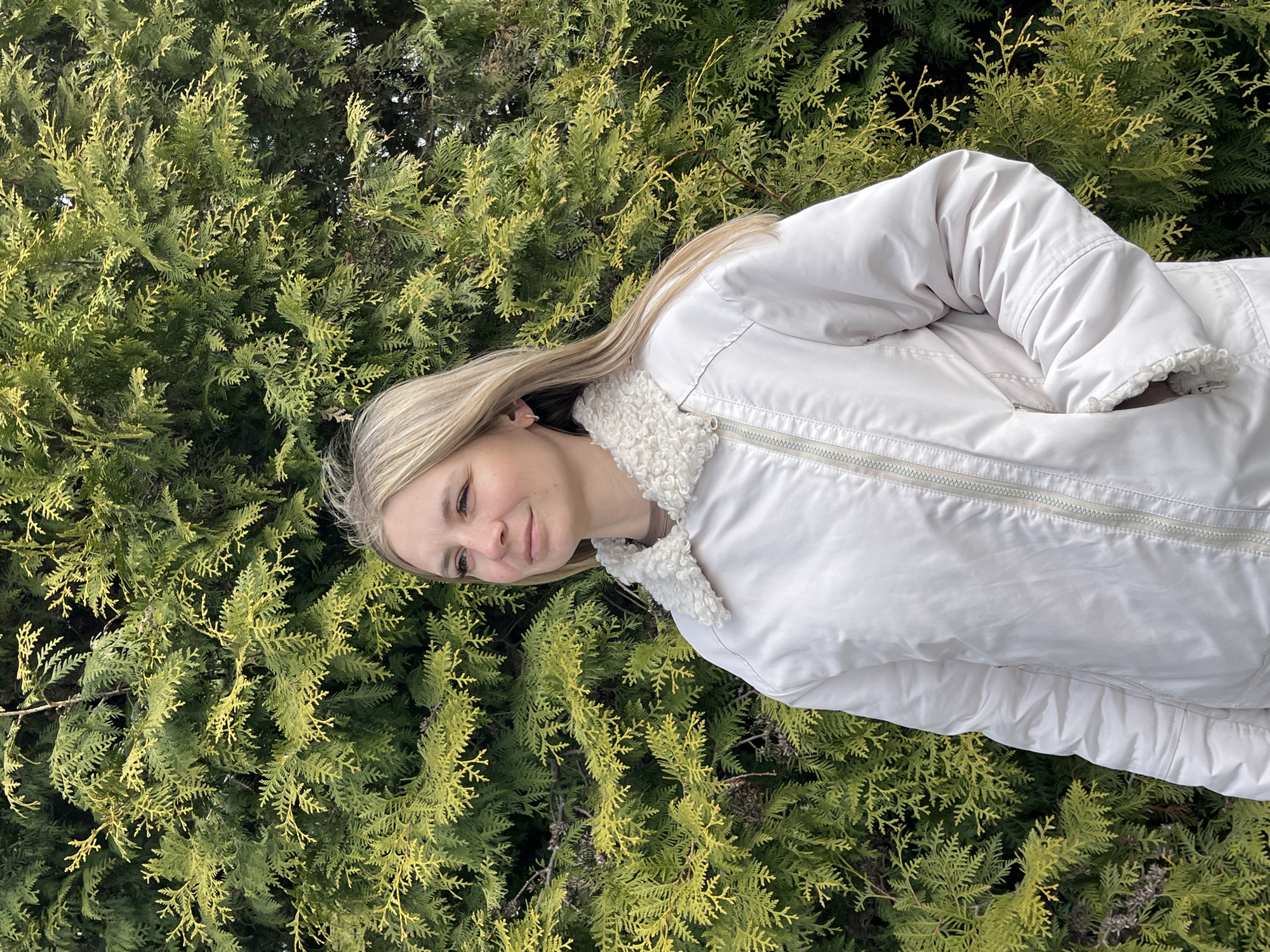 A young, fair-skinned woman with light blonde hair is standing in front of a dense, green shrub with yellow highlights. She is wearing a light-colored jacket with a fluffy, white collar. The location is likely Chaikovsky, Russia, based on the image request. There are no observable actions or interactions. The focus is on the woman and the surrounding vegetation.