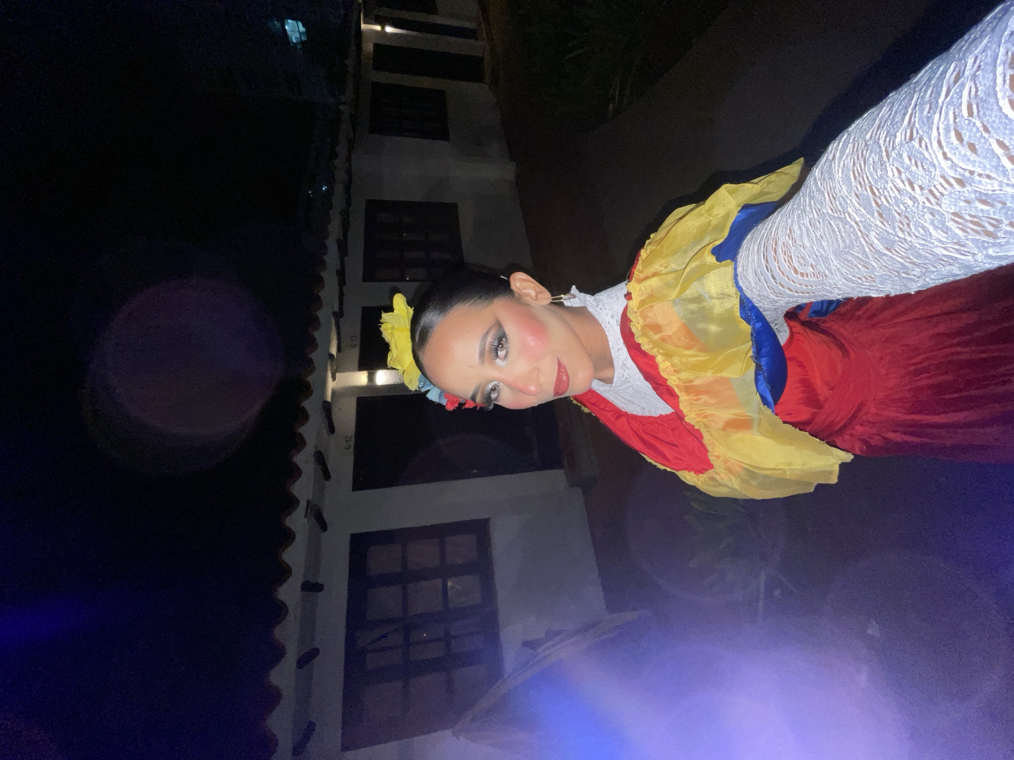 A woman, adorned in traditional Colombian attire featuring a red dress with a yellow and blue ruffled overlay and a floral headpiece, poses for a selfie outdoors at night. The background consists of a row of single-story buildings with tiled roofs, possibly motel rooms, with visible signage indicating room numbers like "6-12" and "6-13". The woman's makeup is dramatic, with bold eye shadow and lipstick. She wears a white lace glove on her right arm, which is extended toward the camera. A blue light flare is visible on the left side of the image, and the overall lighting suggests it is dark outside. The architecture of the buildings, with their white walls and dark-framed windows, is consistent with many tourist accommodations found along the coastal areas of Porlamar, Venezuela.