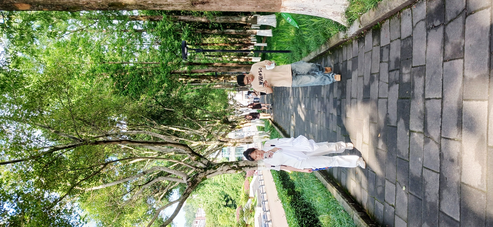 The image shows a partially shaded brick walkway lined with trees and greenery. Two individuals are present: one in white clothing holding a phone, and another in a tan shirt and jeans looking at their phone. Other people are walking further down the path. The location appears to be an outdoor public space in the Chongqing High-tech Zone, China, based on the style of architecture and landscaping.