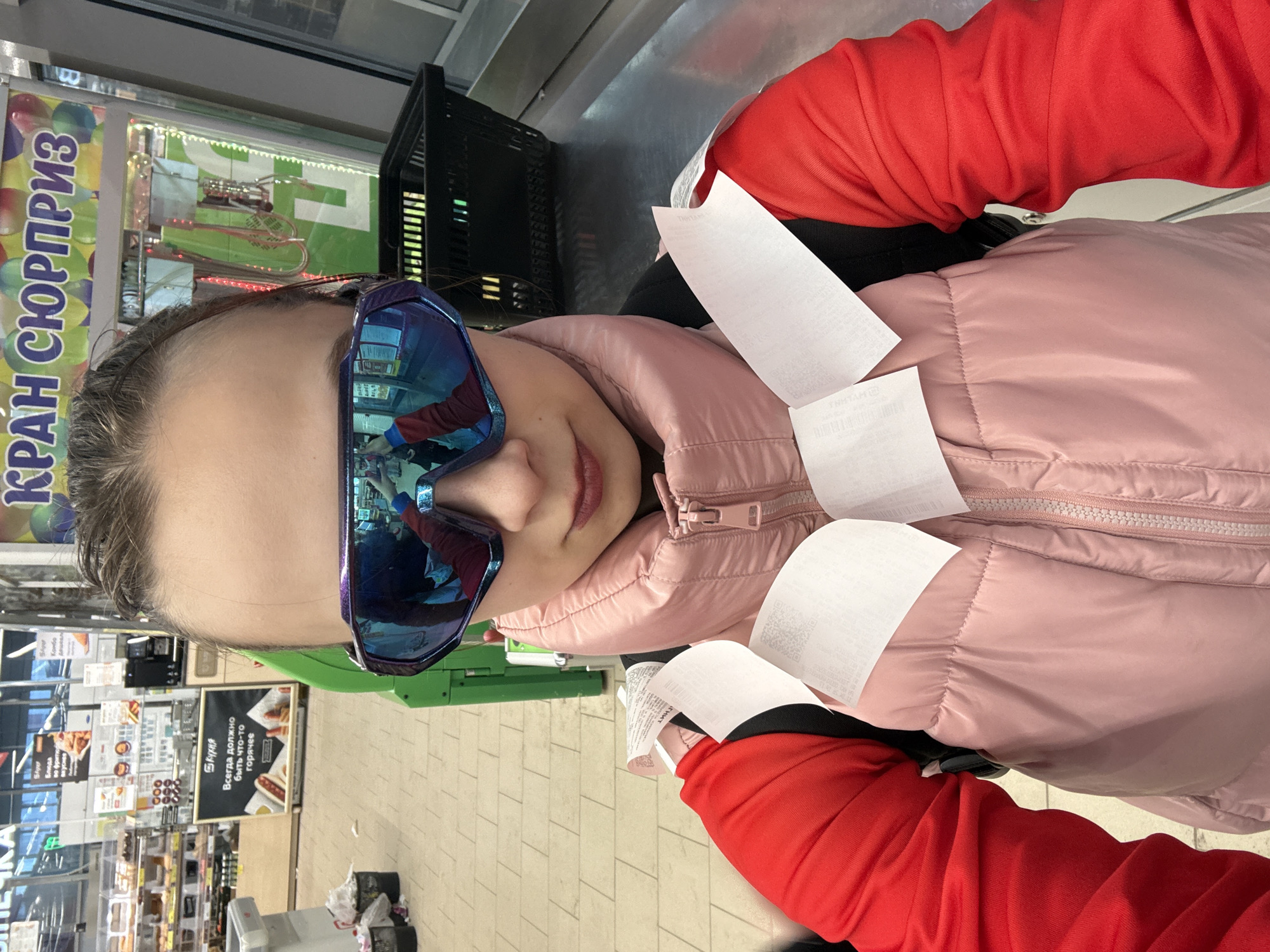 A person wearing blue mirrored sunglasses, a pink puffer jacket, and a red top is standing at a checkout counter in a store. The person is covered in multiple white receipts. The scene includes a black shopping basket behind the person, and the aisle is visible in the background. Text is visible in Russian on nearby signage. Based on the signage and the visible products, the location is a grocery store in Berezniki, Russia.