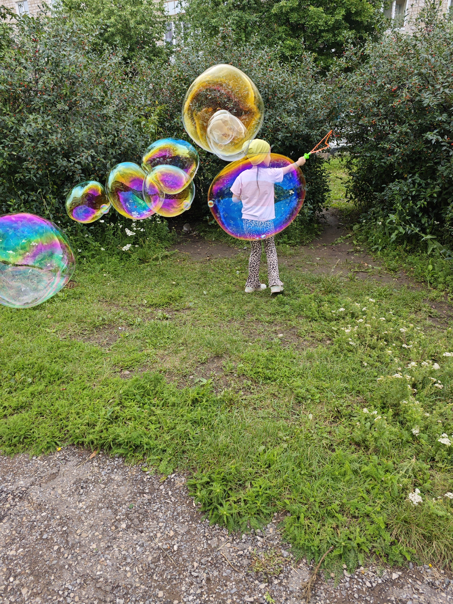 A child wearing a yellow hat, pink t-shirt, and leopard-print pants is blowing large, iridescent bubbles with a green and orange wand. The child stands on a grassy path between two dense green bushes. Several large bubbles are floating in the air around the child, reflecting the surrounding environment. In the background, a brick building is visible through the foliage. The scene appears to be outdoors in a green, natural setting within the city of Tujmazy, Russia.