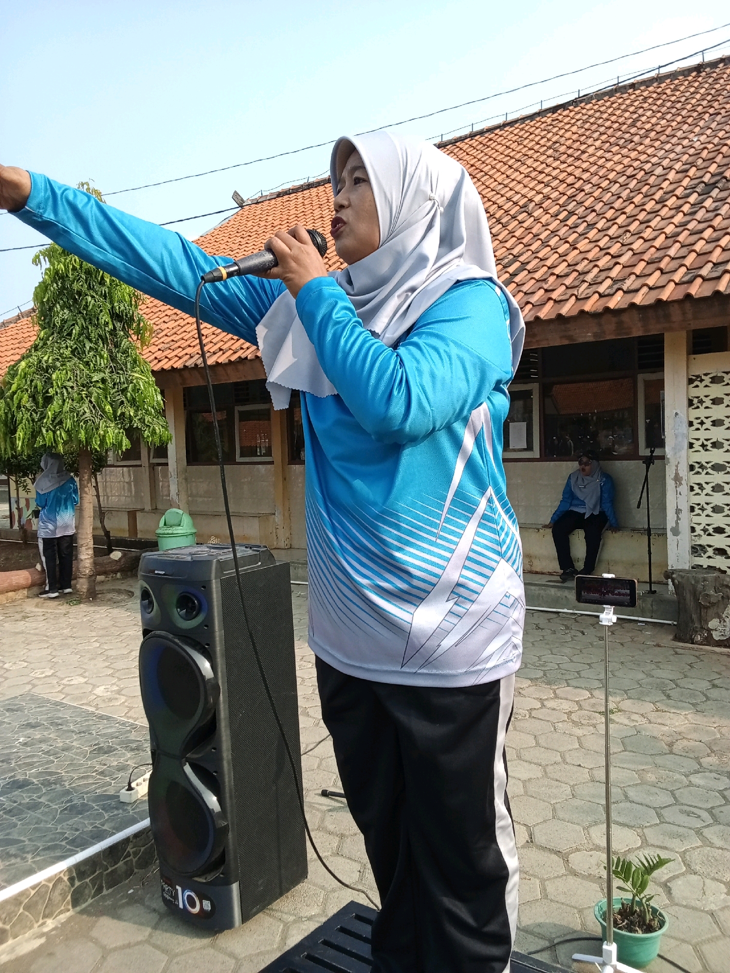 In Tengki, Indonesia, a woman wearing a light blue athletic shirt with white stripes and a light grey hijab, is holding a black microphone and pointing her left arm forward. She is standing next to a large black speaker with blue and red lights. In the background, there is a building with an orange tiled roof and several windows. Another woman in a similar outfit is sitting on a ledge in front of the building. A tree is located to the left of the main subject, and a green trash can is visible behind the speaker. The ground is paved with hexagonal tiles. A small plant in a green pot is on a selfie stick in the foreground.