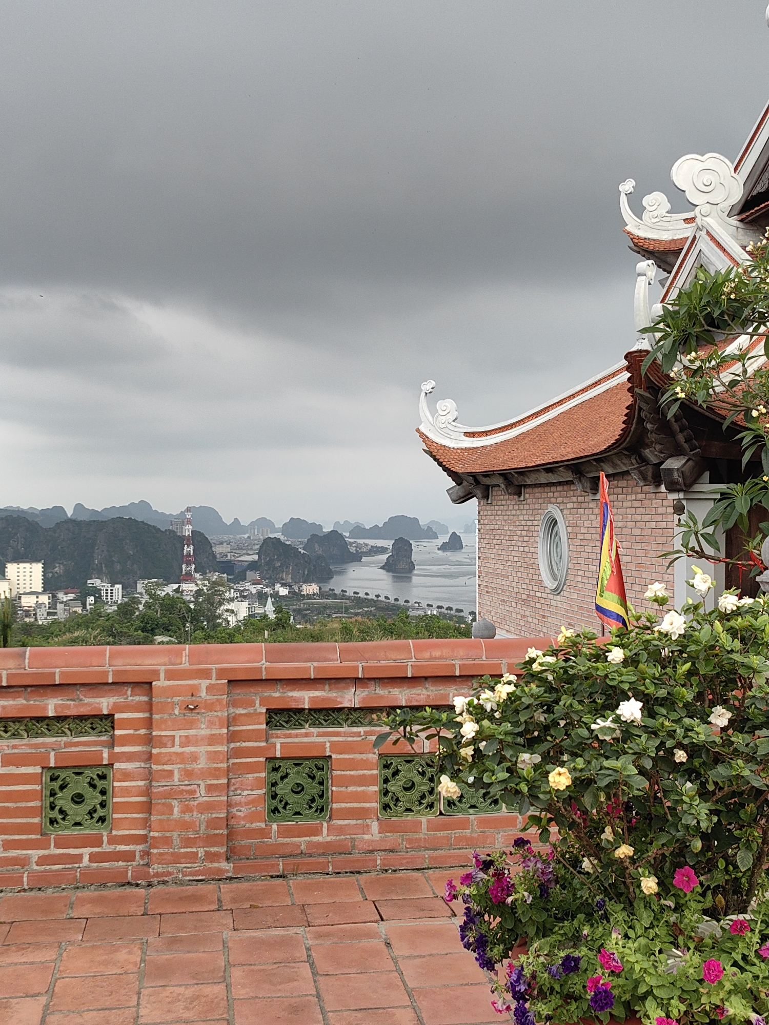The image shows a view from an elevated brick terrace in Ha Lam Ward, Vietnam. A red brick wall with decorative green inserts runs along the foreground. To the right, part of a building with a curved red tile roof and ornamental white trim is visible. A flag is hanging from the building's brick facade. A large bush with white and yellow flowers, and a pot containing purple and pink petunias, stand in front of the wall and building. In the background, a cityscape is visible, with buildings and a red and white communications tower. Beyond the city, a bay with limestone karsts scattered throughout the water stretches into the distance under a cloudy, overcast sky.