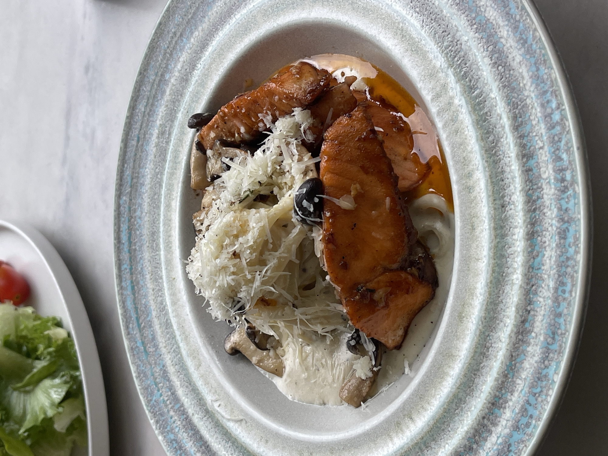 The image depicts a plate of food, likely pasta, topped with cooked salmon, mushrooms, and grated cheese. There appear to be two types of sauce. A partial view of a salad is visible on another plate to the left. The photograph was taken indoors in Da Lat, Vietnam.