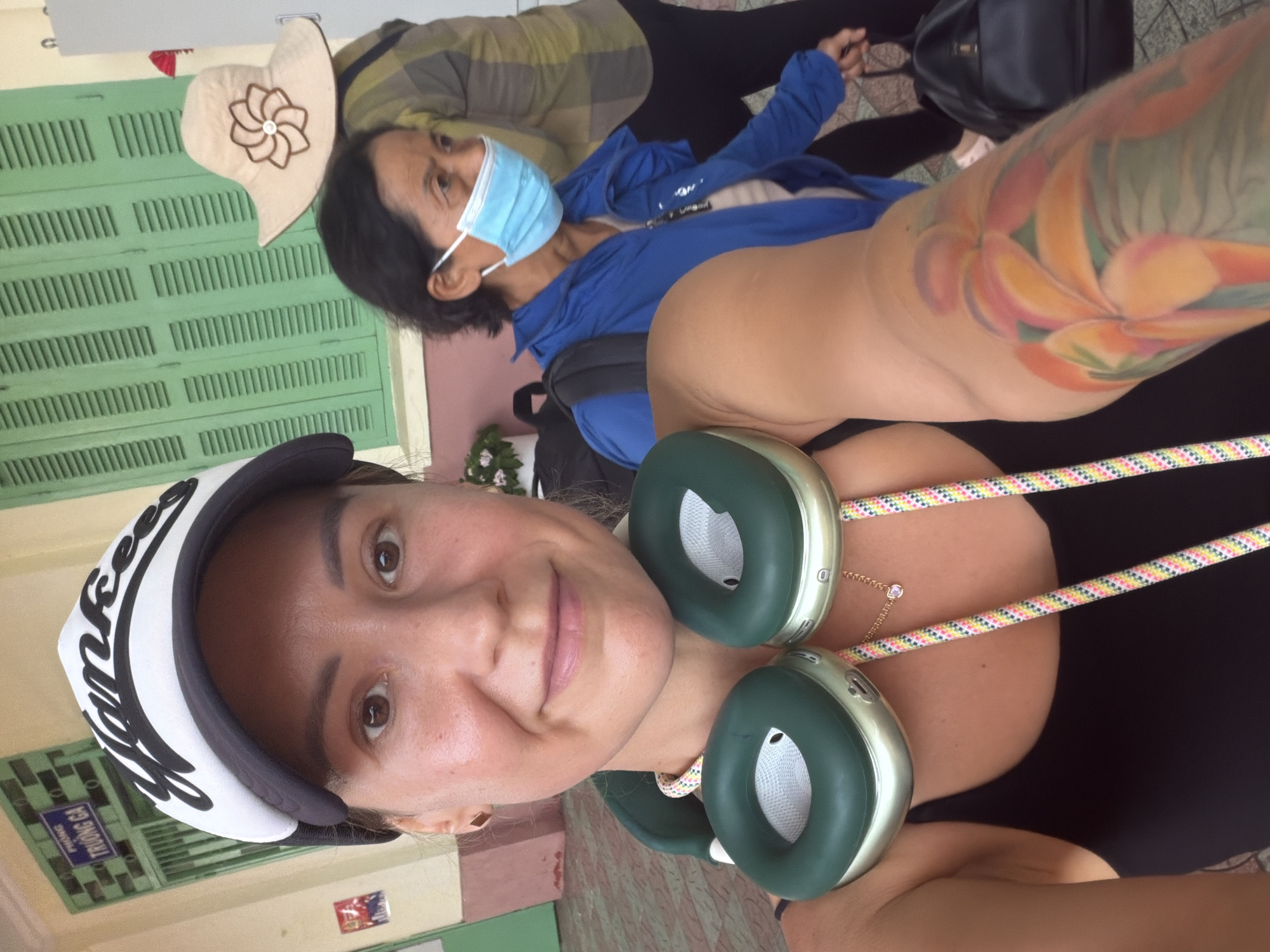 The image shows a woman wearing a black tank top, headphones, and a white and black baseball cap. She has a large floral tattoo on her right arm. Behind her, an older woman with dark hair, wearing a light blue mask and a blue jacket, is walking away from the camera. In the background, there is a green louvered window, a sign with Vietnamese text, and a woman in a plaid shirt and a straw hat. The environment appears to be outdoors, possibly in a city or town. The sign in the background reads "PHUONG TRUONG GA," which translates to "GA Station Area" in Vietnamese. This suggests the location is near a train station in Tay Nha Trang Ward, Vietnam.
