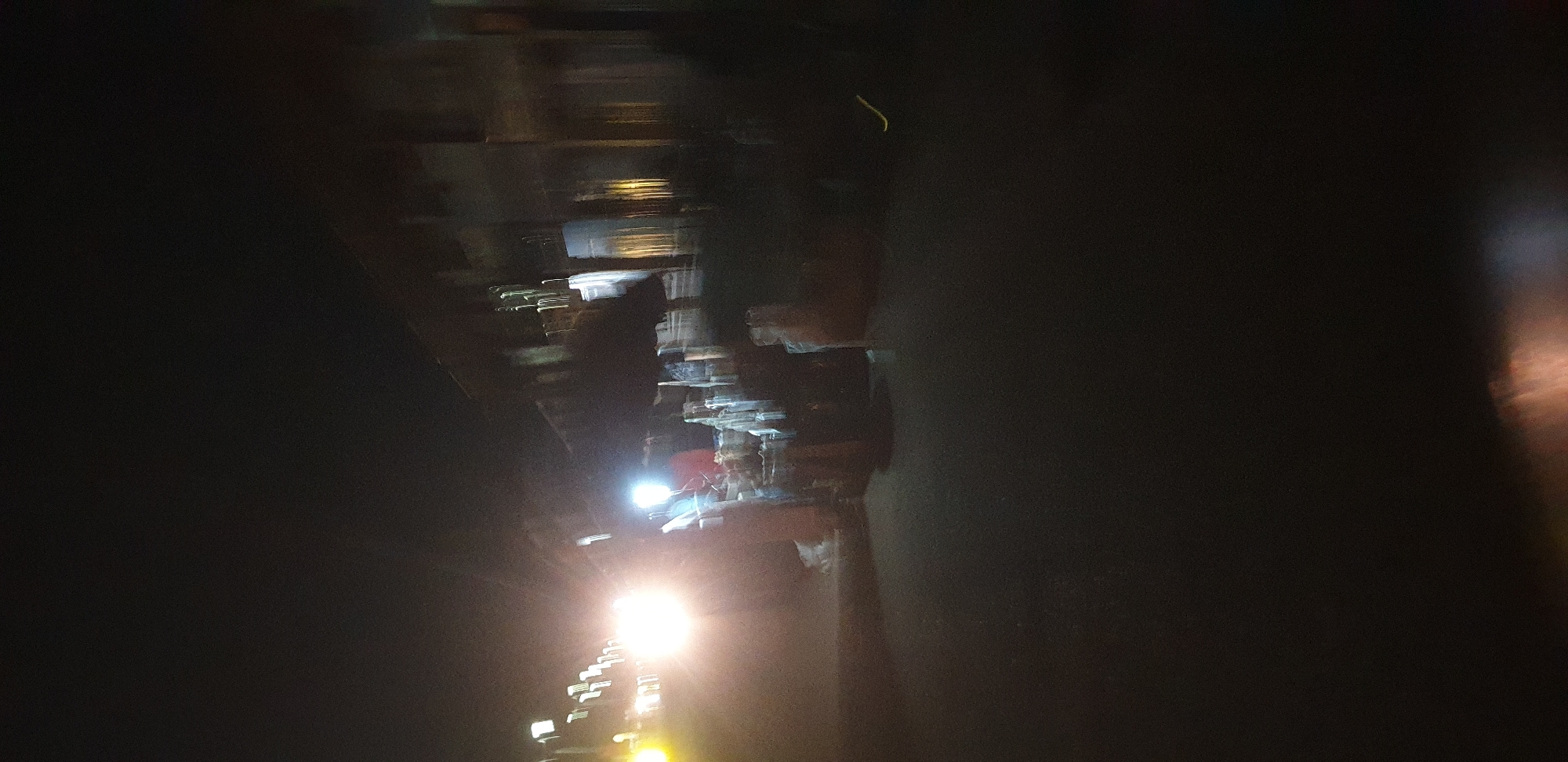 This image depicts a nighttime scene on a street in Sabon Tasha, Nigeria. The environment is dark, with strong artificial light sources dominating the left and central portions of the frame, causing significant glare and lens flare.
In the left-center, a prominent, intensely bright, white-yellow light source is present, likely a vehicle headlight, which emits a strong bloom. To its immediate left, multiple smaller, elongated white and yellow light streaks are visible, indicating other light sources, possibly from additional vehicles or distant streetlights, blurred due to motion or long exposure. A secondary, less intense bright white light appears to the right of the primary light source in the mid-ground.
On the right side of the image, the outlines of multi-story buildings are discernible against the dark sky. These structures appear dark and indistinct due to the low light and motion blur, but some windows or interior lights create vertical streaks of white and yellow illumination.
In the mid-ground, extending from the left towards the right, numerous objects are stacked on what appears to be a road or ground surface. These objects are irregular to rectangular in shape, light-colored, and appear to be covered or wrapped, possibly indicating goods or materials. Their precise nature is obscured by blur and low light.
The ground surface in the foreground and mid-ground is dark, consistent with an unlit road or path. No clearly identifiable people or specific actions are discernible within the blurred scene. The overall impression is one of movement or a long exposure photograph taken at night, characterized by bright light sources and indistinct surroundings.