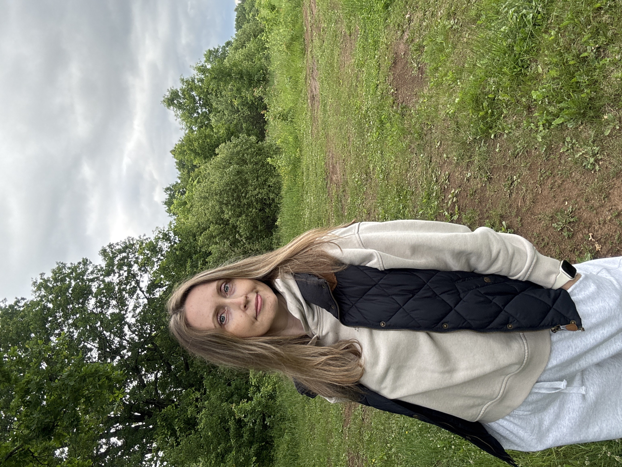 The image depicts an adult female standing outdoors in a grassy area in the city of Chaikovsky, Russia. She is positioned slightly right of the frame's center, angled towards the right, and looking directly at the camera with a subtle smile. She has long, light brown hair that falls over her shoulders. She is wearing a light beige hooded sweatshirt, over which she has donned a dark blue quilted sleeveless vest with brass snap buttons and brown leather-like trim on the collar. Her lower body is clad in light gray sweatpants with a visible drawstring. A white smartwatch is present on her left wrist, and her left hand is partially inserted into her pant pocket. The background features a dense expanse of green trees and bushes under an overcast, cloudy sky. The foreground and midground consist of a green grassy field with interspersed patches of bare brown earth, small white flowers, and other wild vegetation. The lighting is soft and diffused, characteristic of an overcast day.