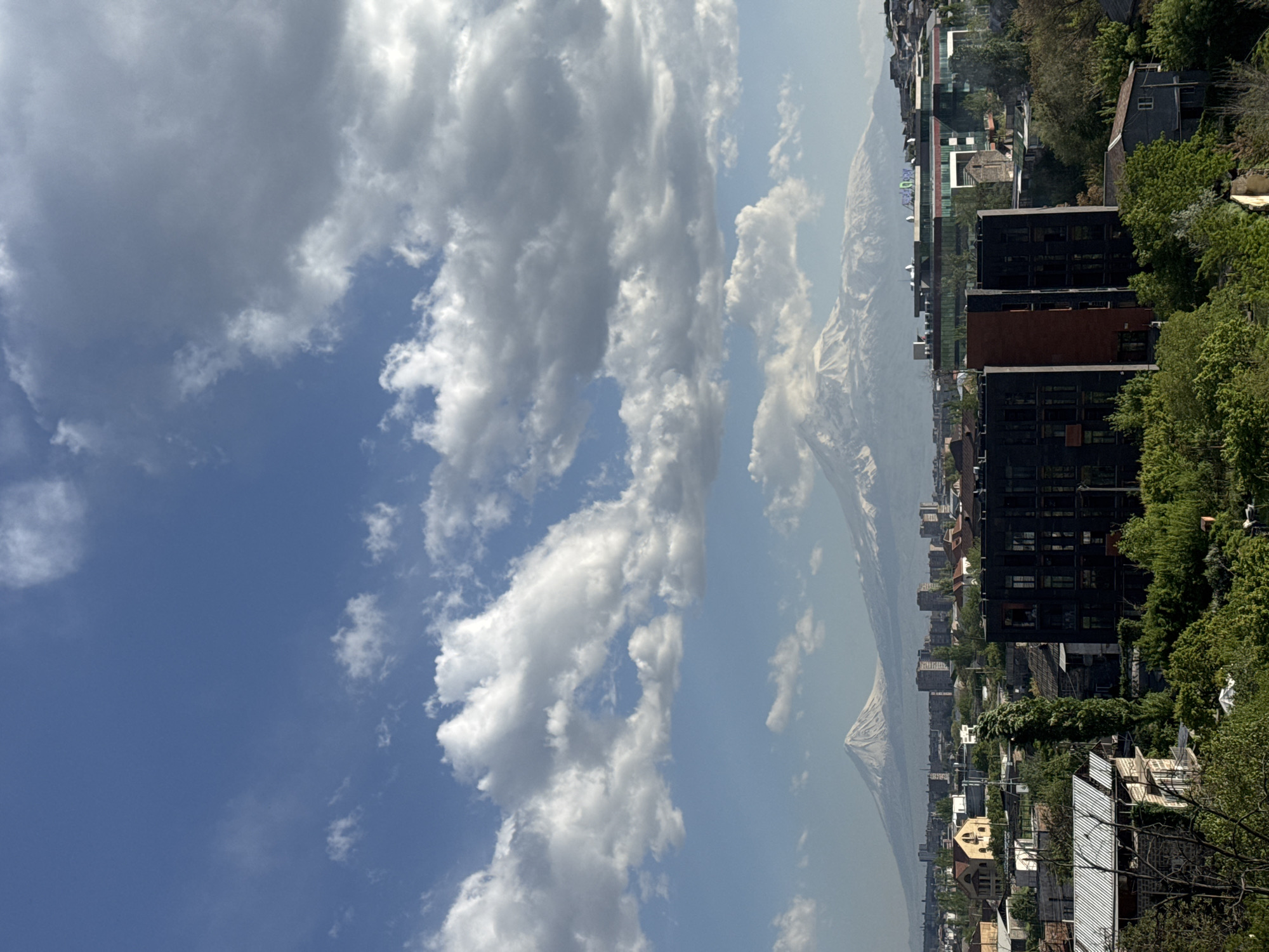 The image shows a view in Yerevan, Armenia. In the foreground, there is a cityscape with buildings and trees. In the background, there is Mount Ararat. The sky is blue with white clouds.