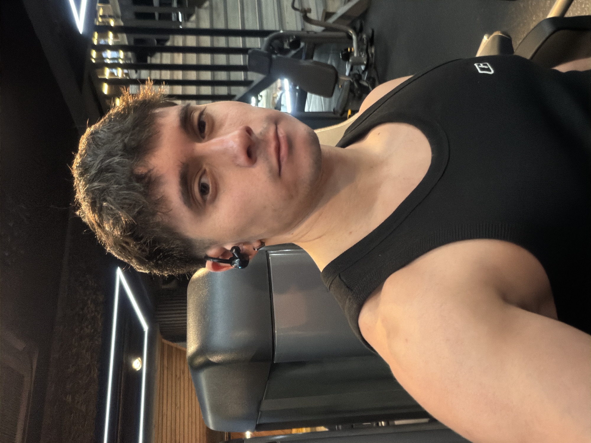 A man is shown in a close-up, slightly angled shot in what appears to be a gym setting. He is wearing a black tank top with a small, white square logo on the upper right chest. The man has short, dark hair and is wearing dark-colored earbuds. In the background, gym equipment and metal bars are visible. The location appears to be a fitness center; however, there are no contextual details that establish this is in Sofia, Bulgaria.