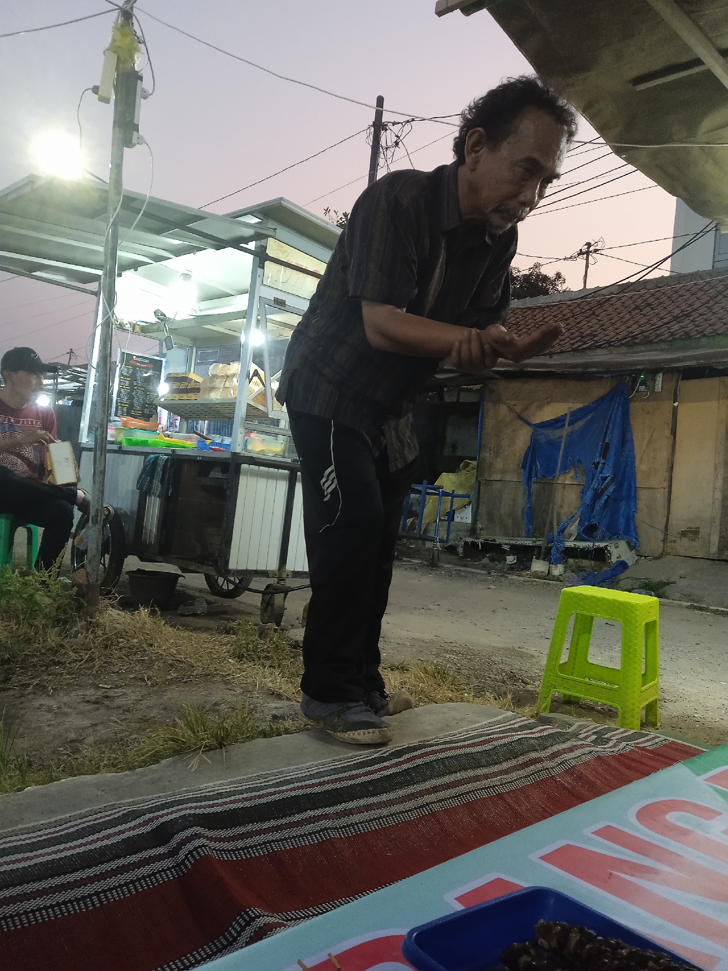 An elderly man with curly hair and a mustache is standing in front of a food cart at dusk in Kaliwungu, Indonesia. He is wearing a dark patterned short-sleeved shirt and dark pants, and he is looking down with his hands clasped together. Behind him, the food cart is illuminated by overhead lights, displaying food items on shelves. To the left, a younger person wearing a red shirt and a cap is sitting on a green stool and holding a piece of bread. The environment includes a dirt ground, dry grass, a concrete path, and a building with a blue tarp hanging from it in the background. A bright green plastic stool is visible on the right side of the frame, and a mat with a striped pattern is laid out on the ground in the foreground.