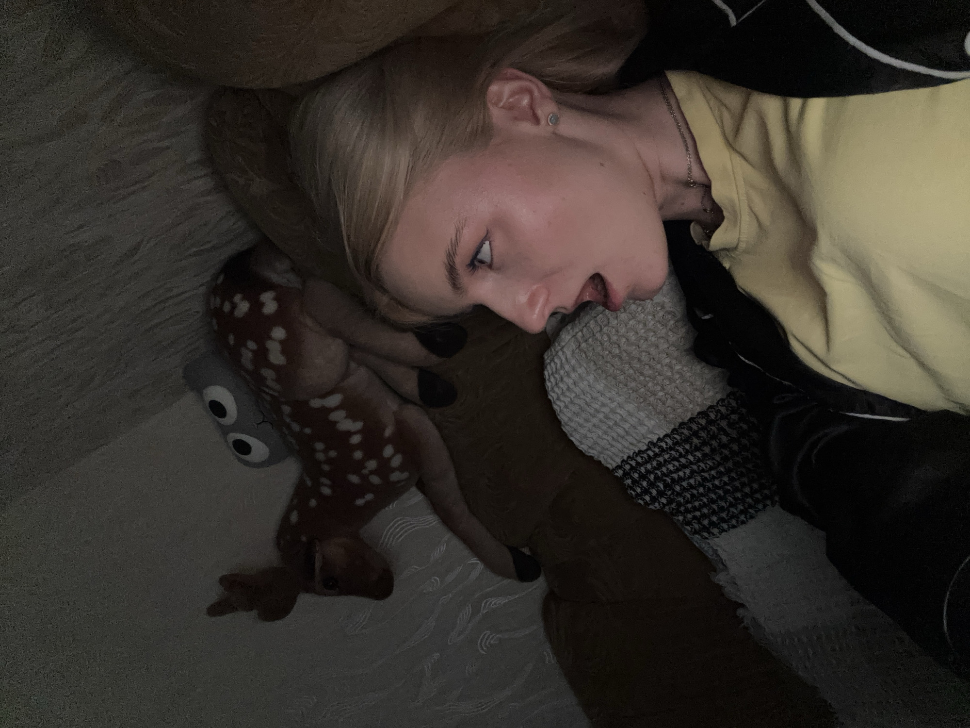 A blonde woman with her mouth open in surprise is lying down in a dimly lit room. She is wearing a yellow t-shirt and a black jacket. Beside her is a plush toy deer with white spots and large cartoon eyes. The woman's head is resting on a brown cushion, and she is partially covered by blankets with black and white patterns. The background shows a textured wall and the corner of a couch. The overall impression is a candid, informal moment.