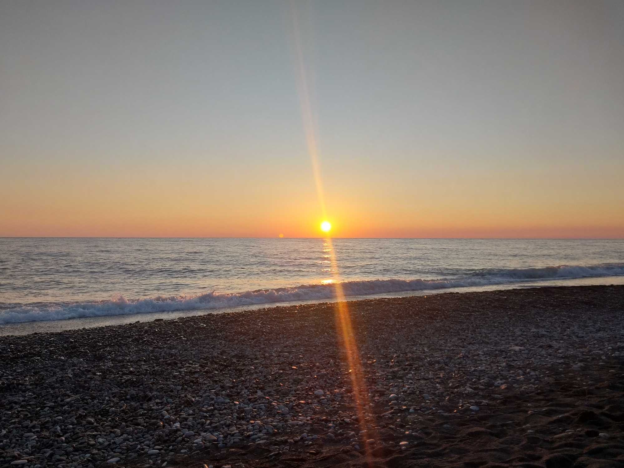 The image displays a serene coastal landscape captured during either sunrise or sunset in Sochi, Russia. The sun, a brilliant yellow-orange orb, is positioned low on the horizon above the sea, emitting a strong sunburst or lens flare that extends towards the viewer and across the water.
The sky exhibits a clear, graduated palette, transitioning from warm oranges and soft yellows near the horizon, illuminated by the sun, to a lighter, delicate blue-grey hue higher in the frame. The sea is calm, with gentle waves breaking into white foam along the shoreline. A shimmering path of sunlight reflects brightly on the water's surface, leading directly from the sun to the beach.
The foreground consists of a dark pebble beach, with small, round stones covering the majority of the visible shore. Some darker sand is also interspersed, particularly closer to the bottom edge of the frame. The pebbles appear somewhat wet and subtly reflect the soft ambient light.
There are no people, animals, or man-made structures visible, emphasizing the untouched natural beauty of the scene. The overall impression is one of peacefulness and tranquility. No visible text is present in the image.