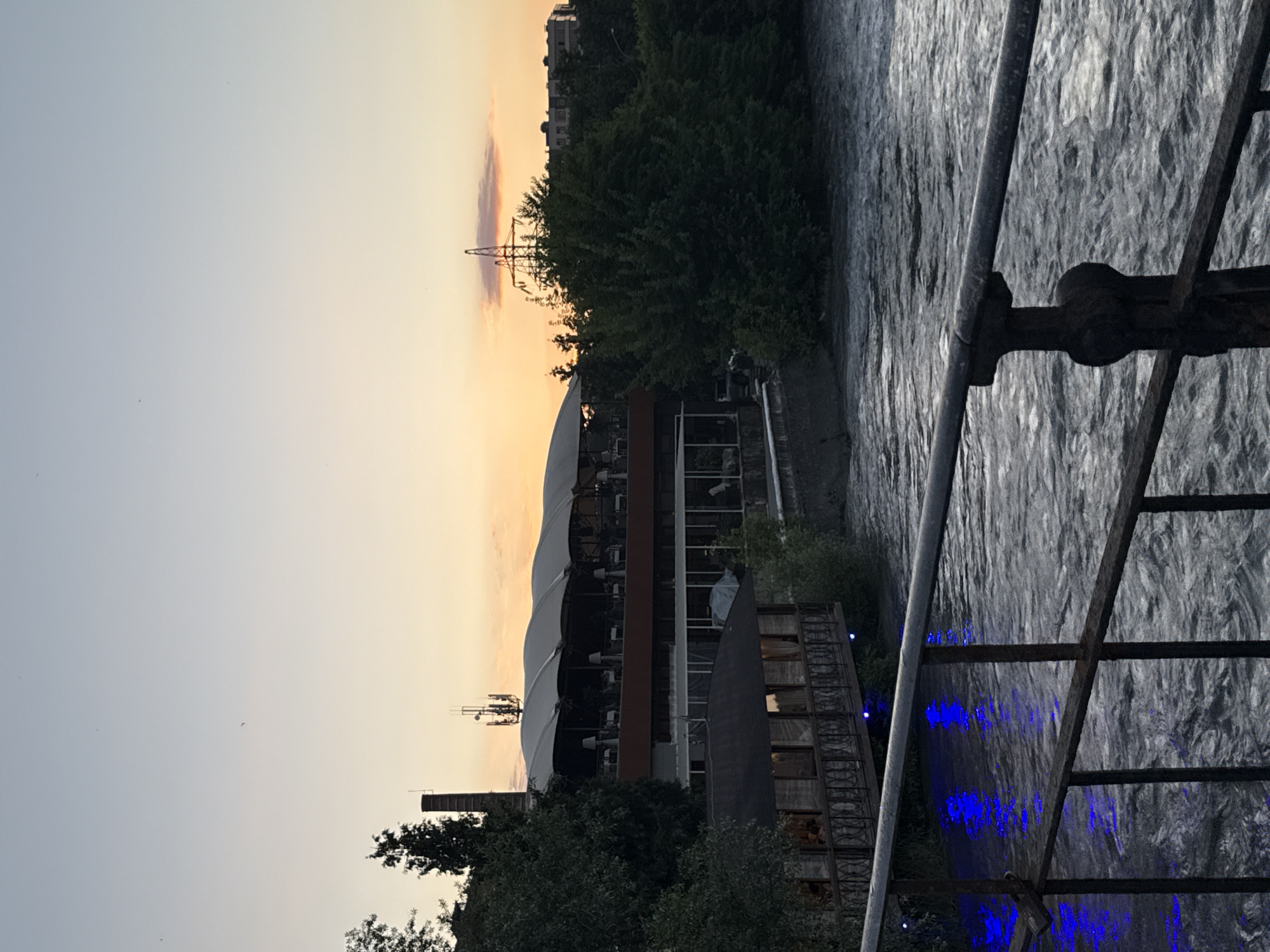 The image depicts a sunset over a river in the city of Askhazan, Russia. A multi-story restaurant with a distinctive white, undulating roof is situated along the riverbank, with tables and chairs visible on its terraces. Below the main restaurant, a smaller, dark-colored wooden structure with a covered walkway and ornate railings is also visible, illuminated by blue lights reflecting in the water. The river itself is wide and appears to have a moderate current. A metal railing in the foreground suggests the viewpoint is from a bridge or walkway overlooking the river. Trees and other buildings are visible on the opposite bank, along with power lines and a tall industrial chimney. The sky is a gradient of pale blue to orange, indicating the setting sun. No people are clearly discernible in the image.