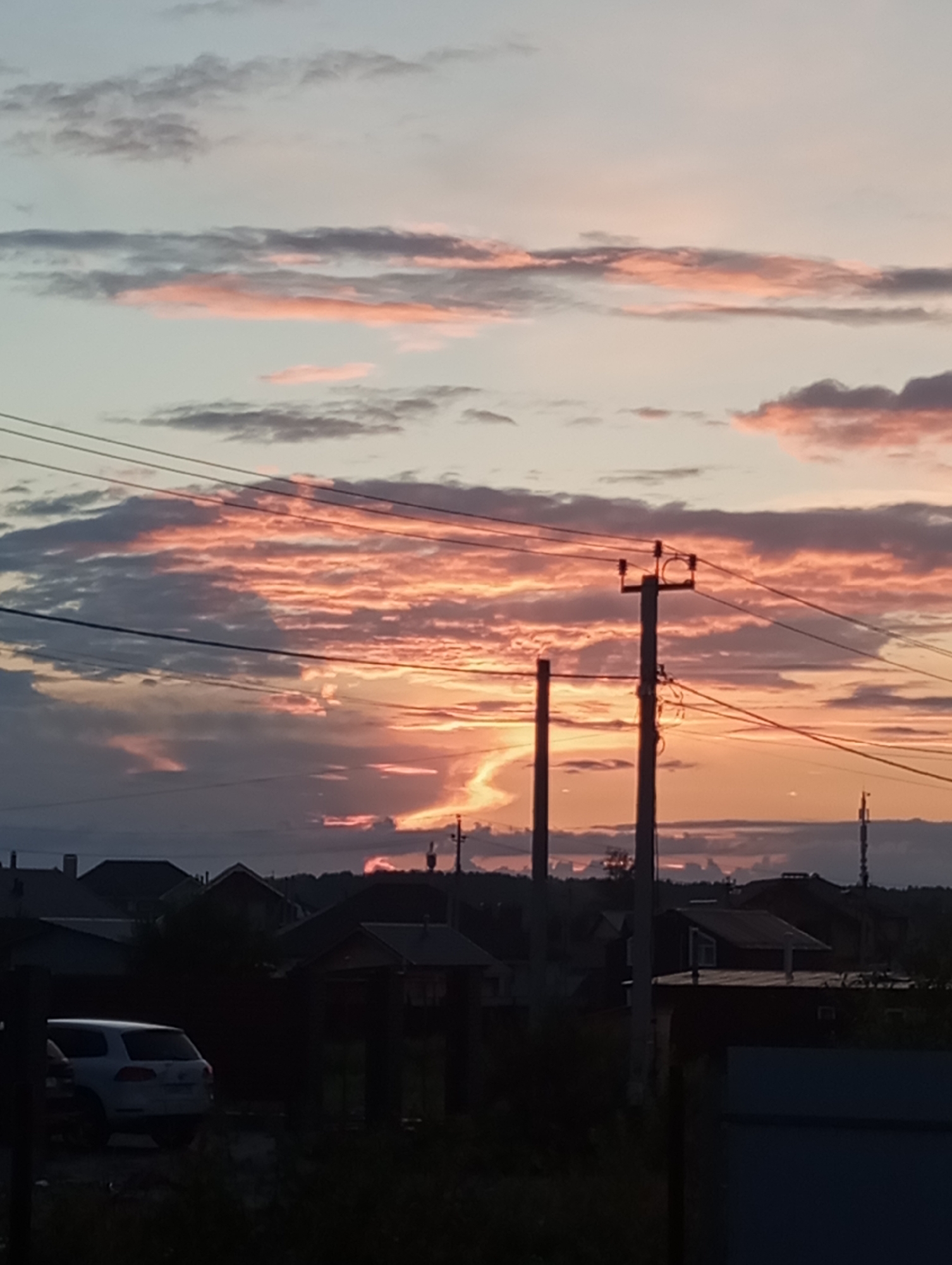 The image depicts a rural or suburban landscape at dusk, likely in or near Verkhnyaya Pyshma, Russia, characterized by a dramatic sunset. The sky is filled with clouds illuminated in shades of orange, pink, and gray, with streaks of sunlight breaking through. In the foreground, power lines traverse the frame, supported by wooden poles. A white SUV is visible on the left, parked near a dark building. The background shows a silhouette of houses with pitched roofs against the darkening sky, and a treeline in the far distance. The overall scene conveys a sense of the end of the day in a residential area.