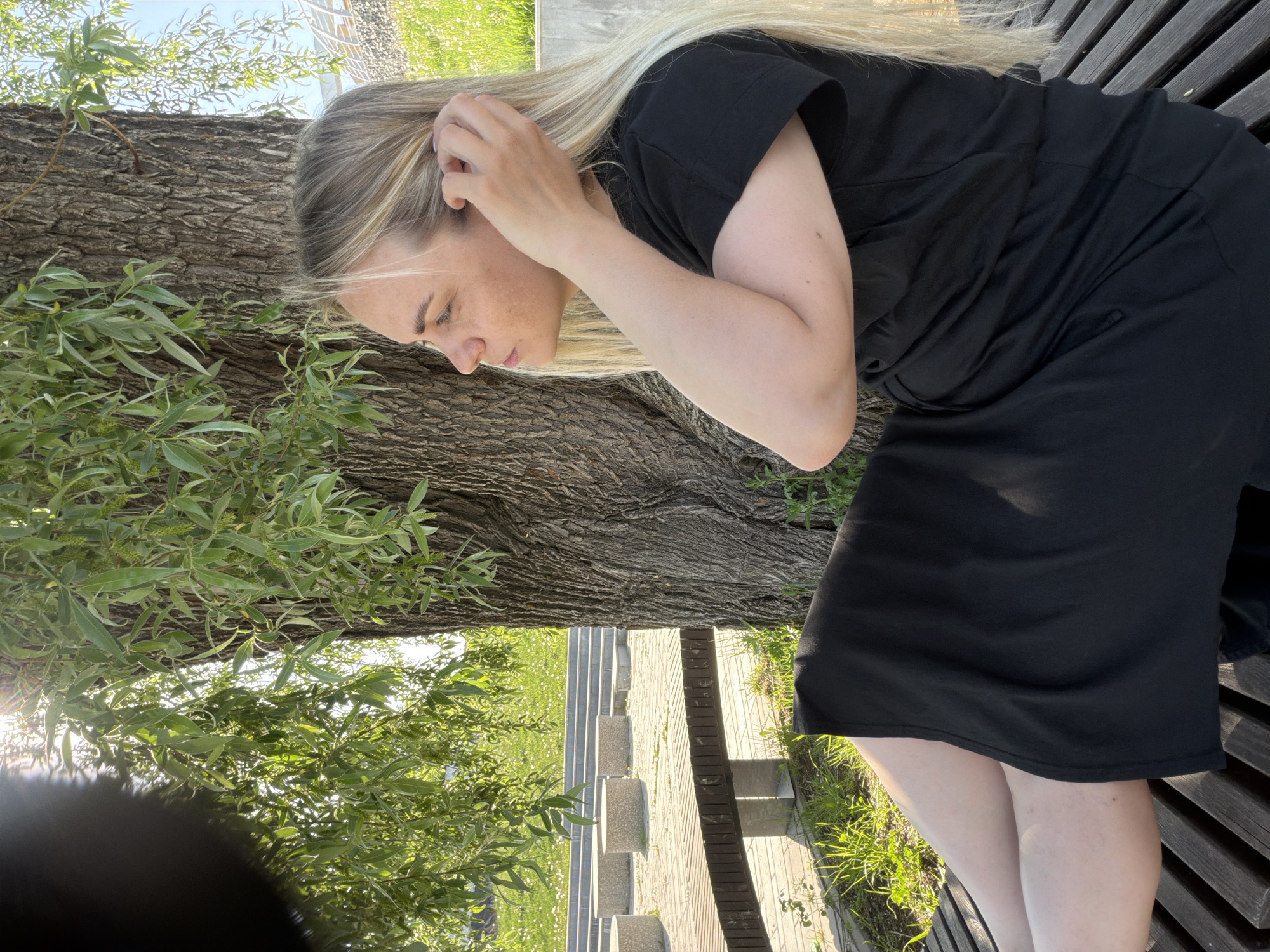 A woman with long blonde hair is sitting on a wooden bench outdoors. She is wearing a black dress. Her right hand is touching her hair near her ear. The bench is next to a large tree with green leaves. Behind the bench is a stone pathway with several short, square stone pillars. Grass and other greenery are visible in the background. The location is Votkinsk, Russia.