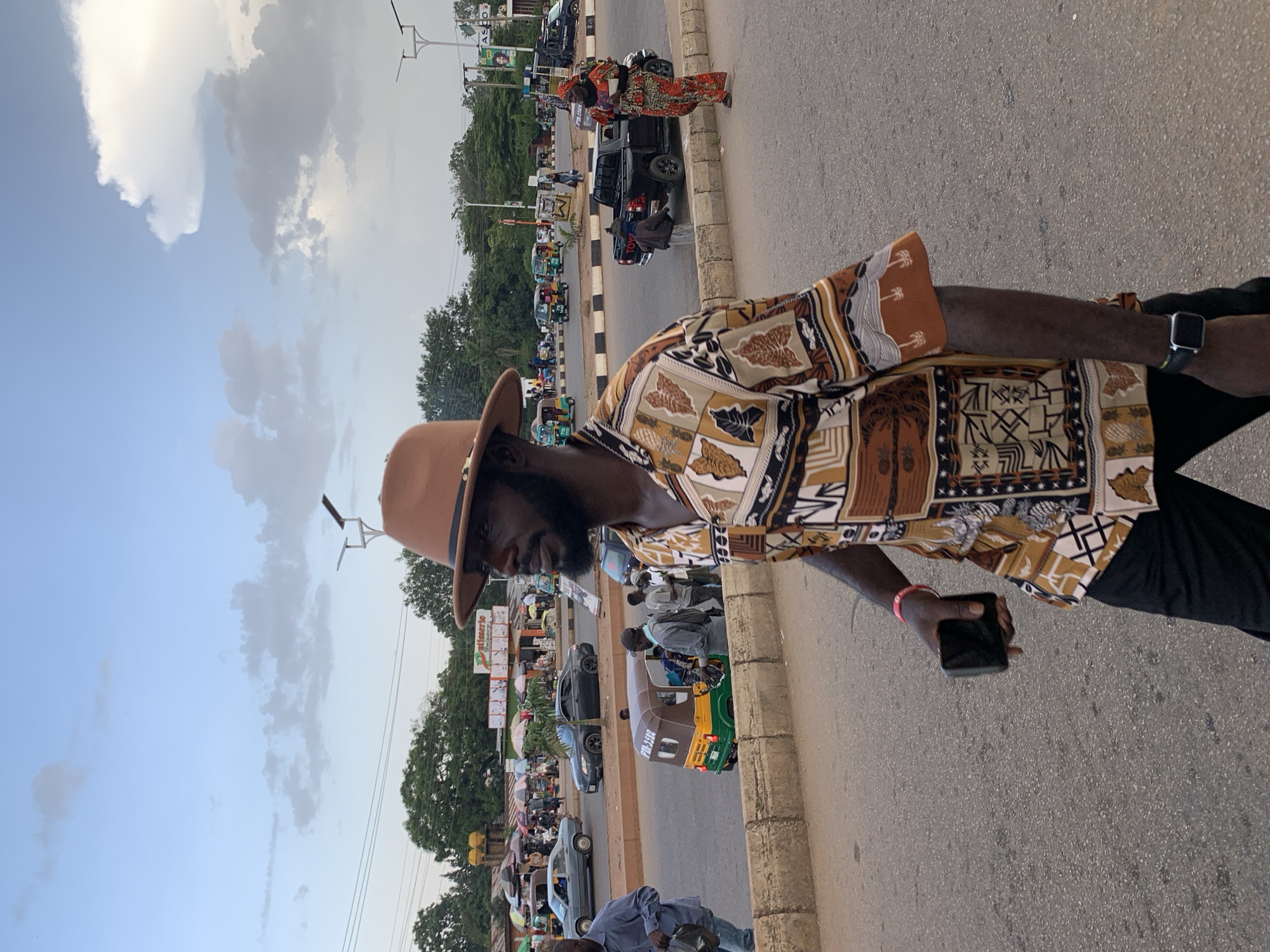 The image captures a street scene in the city of Jos, Nigeria, likely near a busy intersection or market area, with a focus on a man in the foreground. He is walking towards the right of the frame, wearing a brown fedora hat, a short-sleeved shirt with a vibrant, geometric African print in shades of brown, tan, and white, and black trousers. He holds a dark-colored smartphone in his left hand and wears a smartwatch on his right wrist.
The background reveals a typical urban environment in Jos. Cars, including a grey sedan and a black pickup truck, are visible on the road. A colorful three-wheeled auto-rickshaw, commonly known as a Keke Napep, is also present, with passengers visible inside and around it. In the distance, a marketplace or collection of shops lines the street, with umbrellas providing shade over stalls. People are seen walking and interacting in the background, suggesting a lively and active commercial area. The sky above is partly cloudy, with a mix of light and darker grey clouds against a pale blue sky, indicating either late afternoon or early evening. The overall scene depicts everyday life and commerce in Jos, Nigeria.