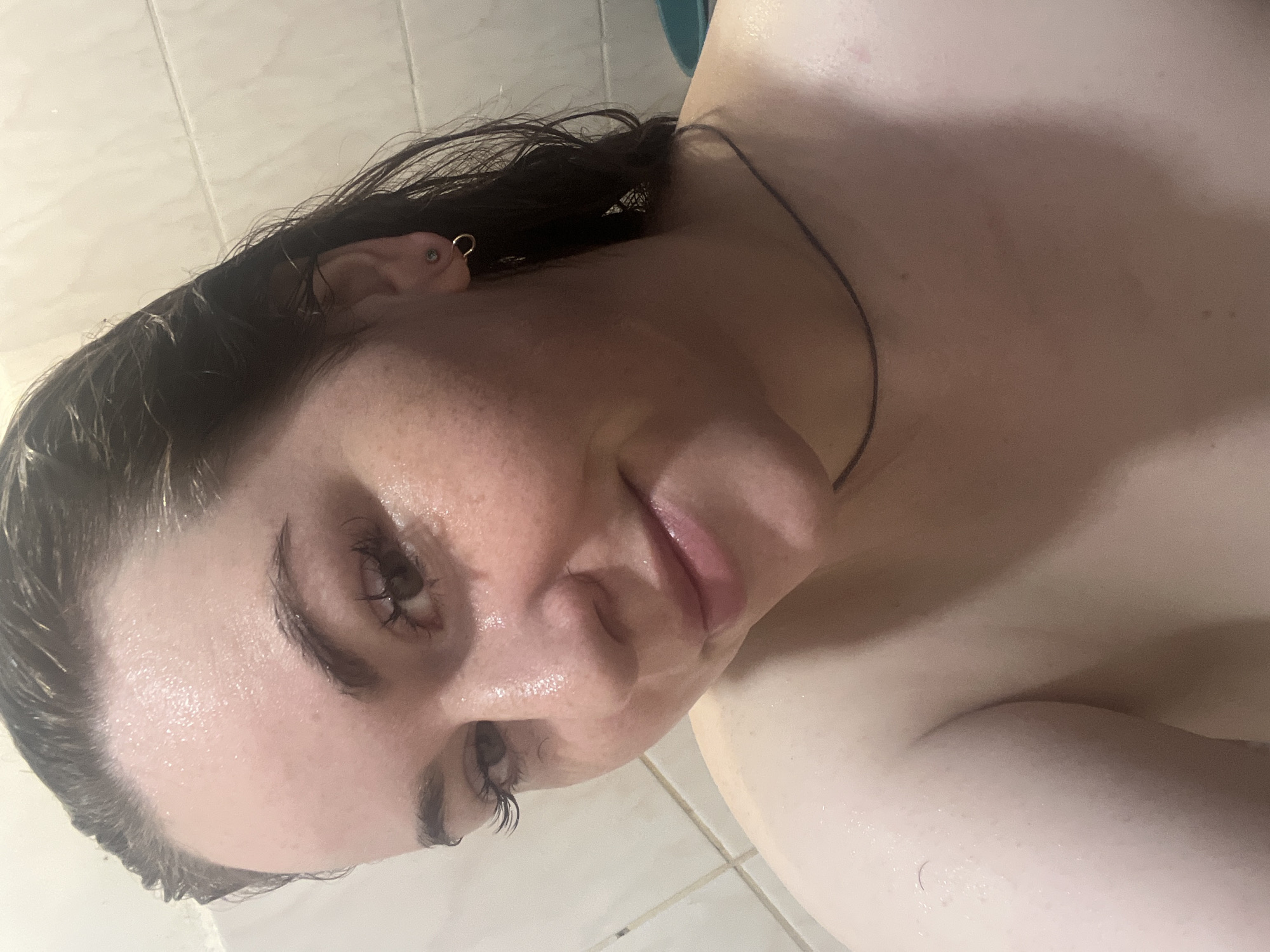 This close-up image features an adult individual, likely female, with dark, wet hair and glistening skin, suggesting she is in a shower or a humid environment. Her face is angled towards the camera, with a neutral to subtly relaxed expression. She has a small, metallic ring earring in her right ear, along with a stud. A thin, dark cord or necklace is visible around her neck.
The background consists of light-colored, tiled walls, indicative of a bathroom or shower stall. The wet appearance of the subject's hair and skin, combined with the tiled setting, points to the activity taking place being a shower or directly after one. The image appears to be a self-portrait. The setting is an indoor bathroom in Chaikovsky, Russia. There is no visible text.