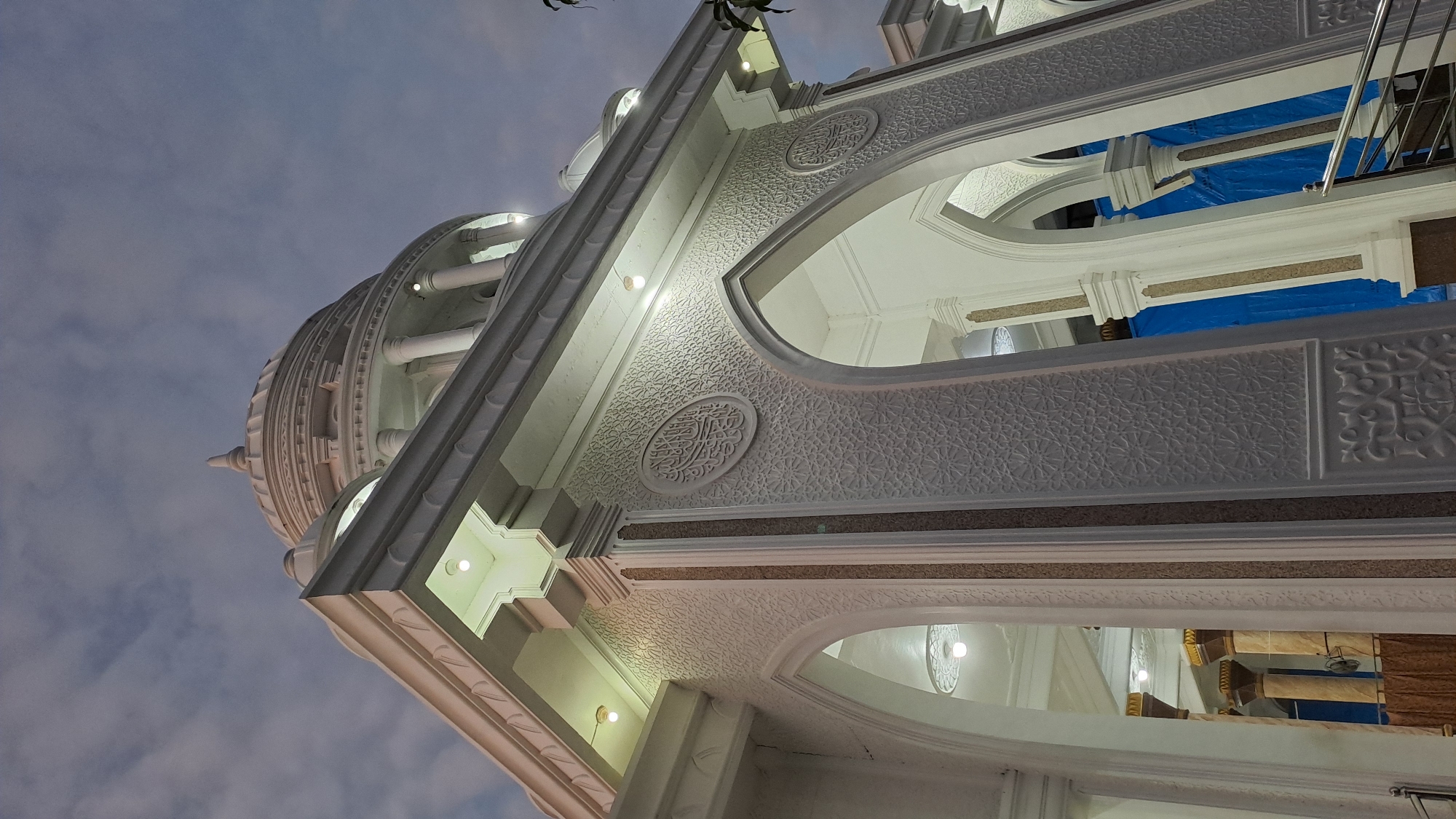 This is a low-angle, full-shot, outdoor photograph of the exterior of a mosque in Lhokseumawe Teungoh, Indonesia, captured at dusk. The mosque is predominantly white with ornate architectural details. A prominent dome is visible at the top of the structure, surrounded by columns and decorative moldings. The facade features arched doorways and intricate relief carvings on its walls. Several lights are illuminated on the exterior, casting a warm glow against the twilight sky, which is filled with diffused clouds. Inside the arches, the blue tarping suggests ongoing construction or renovation. The scene appears serene and majestic, highlighting the mosque's grandeur against the fading light. There are no people visible in the frame.