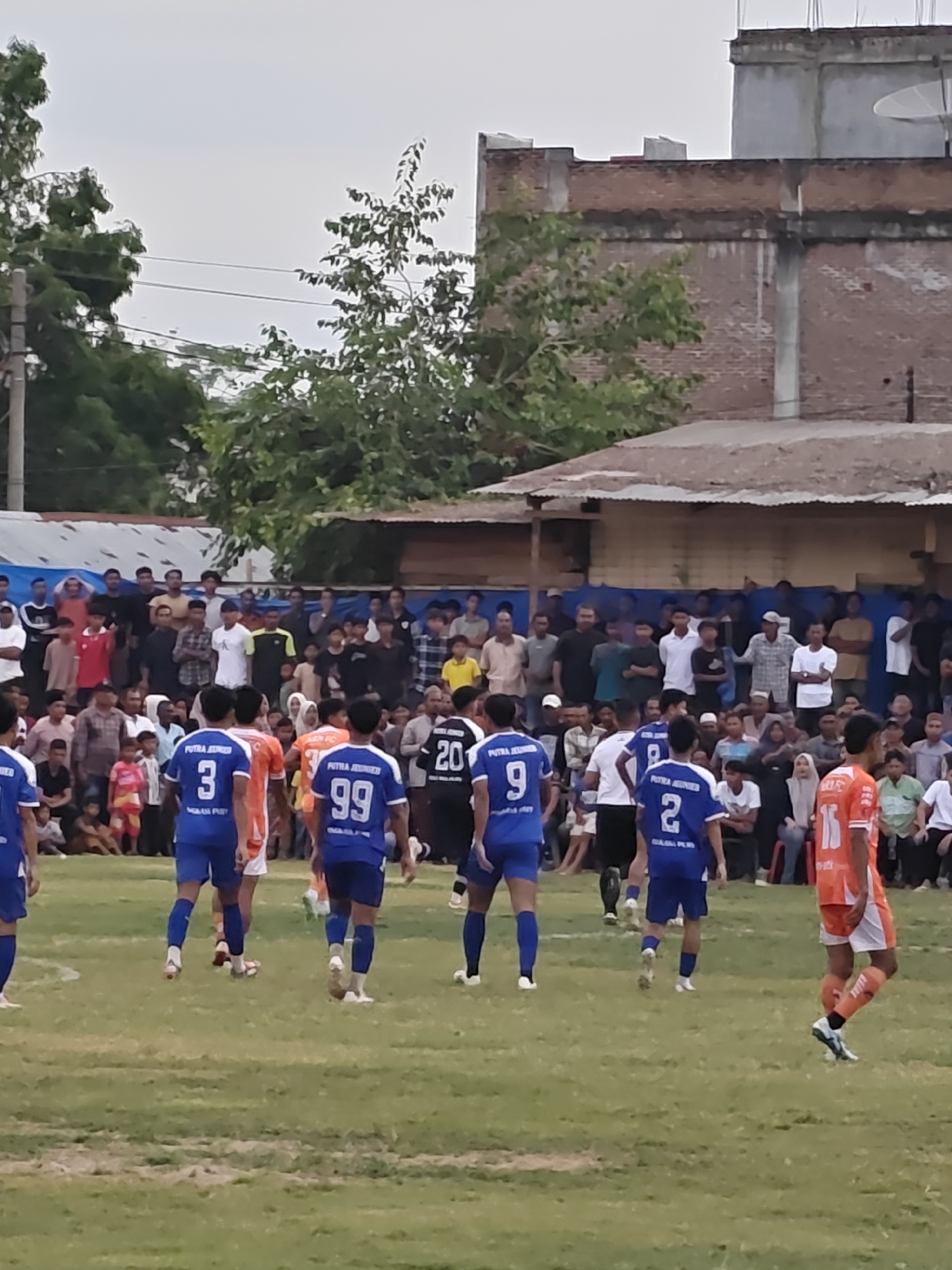 This is a medium shot photograph taken outdoors during the daytime, likely in the late afternoon due to the soft lighting. The scene depicts a football match in the village of Desa Meunasah Blang, Indonesia.
In the foreground, several male football players are visible on a grassy field. Some are wearing blue jerseys with numbers, such as "3", "9", and "99", and blue shorts. Another player is wearing an orange jersey with the number "15" and orange socks. Their stances suggest they are actively participating in or observing the game. Their faces are mostly obscured or turned away from the camera.
Behind the players, a crowd of spectators is gathered. They are of various ages and genders, dressed in casual clothing. Some are standing, while others appear to be seated or leaning against structures. The crowd forms a dense backdrop, suggesting an event with significant local interest.
In the background, there are simple, weathered buildings, possibly houses or community structures, with corrugated metal roofs. Trees with lush green foliage are also present, contributing to the rural atmosphere of the location. Power lines and poles are visible above the trees, indicating the presence of basic infrastructure. The sky is overcast, with a muted grey tone. The overall environment appears to be a typical village setting in Indonesia.