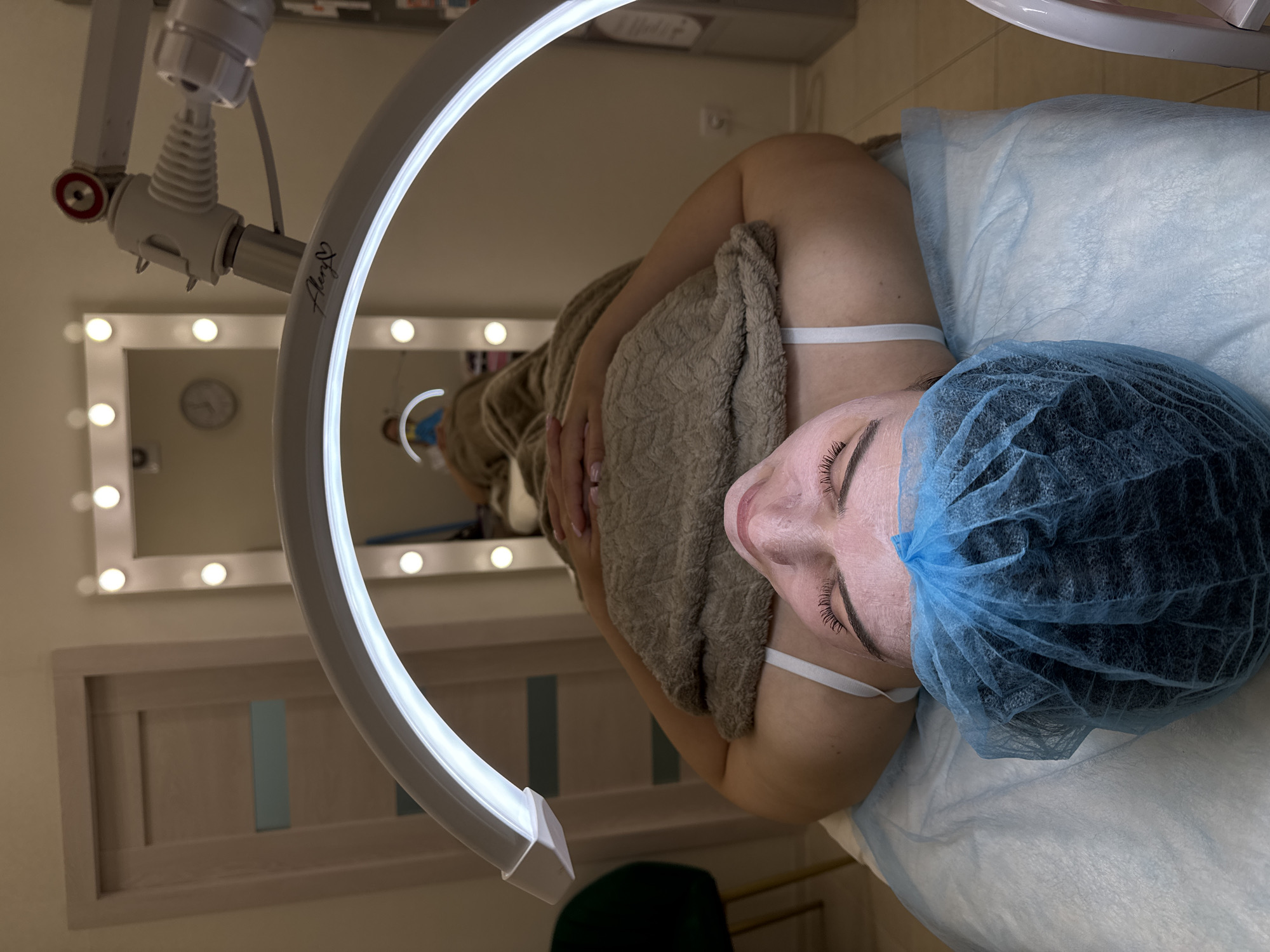 The image depicts an indoor scene within what appears to be a beauty salon or treatment room. The main subject is a person, likely female, lying horizontally on a treatment bed, seen from an overhead perspective. Their eyes are closed, and their face is covered with a light-colored facial product or mask. They wear a blue disposable hairnet to keep their hair contained, and a white strap from an undergarment is visible on their shoulder. A textured, light brown towel covers their torso. The person is lying on a light blue disposable sheet that covers the treatment bed.
Dominating the foreground and positioned over the person's head is a large, illuminated semi-circular LED ring light, suspended by an adjustable metallic arm. On this arm, a small, stylized logo or signature is visible, reading "Anya ♡".
In the background, a large rectangular vanity mirror with multiple incandescent bulbs along its frame reflects a portion of the room, including a round clock with a white face. Below the mirror, there is light-colored wooden wall paneling or a shelf with several faint blue vertical accents. A dark green object, possibly part of a chair or cushion, is partially visible in the lower foreground. The scene suggests a beauty or skincare treatment is actively in progress, with the ring light providing optimal illumination for the procedure.