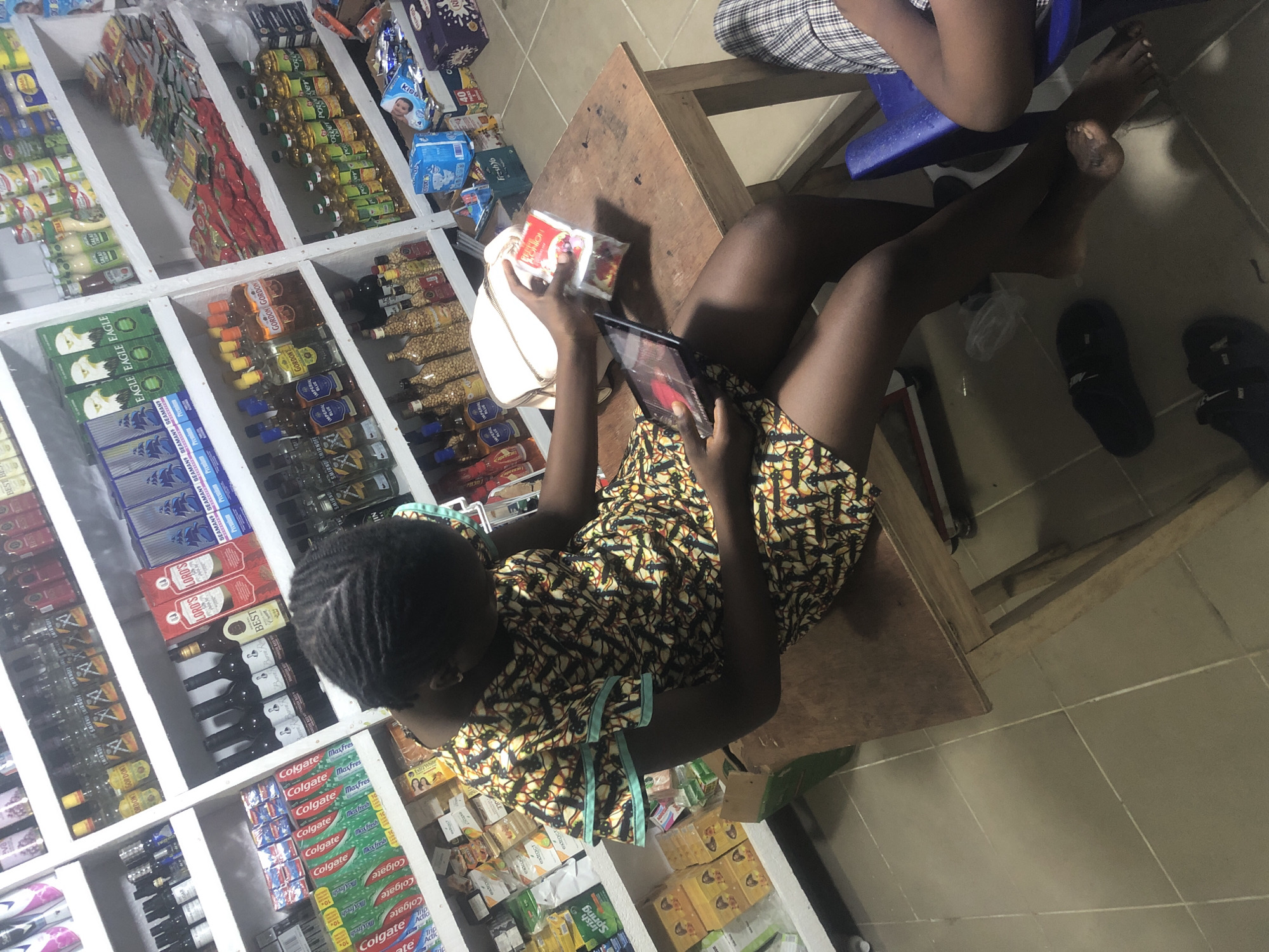 The image depicts the interior of a small convenience store in Ado, Nigeria. A young Black woman is seated at a rustic wooden table, engrossed in a tablet. She is wearing a brightly patterned dress with short, flared sleeves and appears to be examining a small package in her hands. The store is densely stocked with shelves filled with various consumer goods, including bottled beverages, packaged foods, toiletries, and cleaning supplies. The lighting is somewhat dim, suggesting indoor illumination. The floor is tiled. Another person's legs, wearing plaid pants, are visible on the right side of the image, indicating their presence in the store, possibly seated.