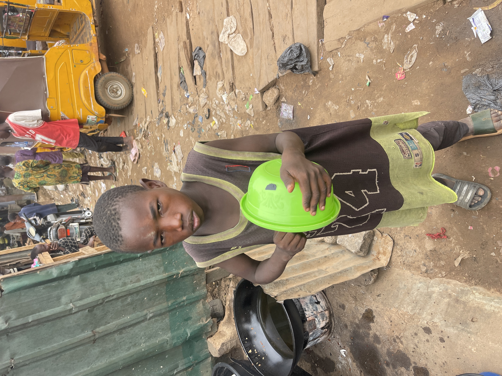 A child stands in the Chikaji, Nigeria neighborhood, holding a green plastic bowl, next to metal cooking pots. The surrounding area is littered with trash and debris, and a yellow vehicle is partially visible. Other individuals are present in the background.