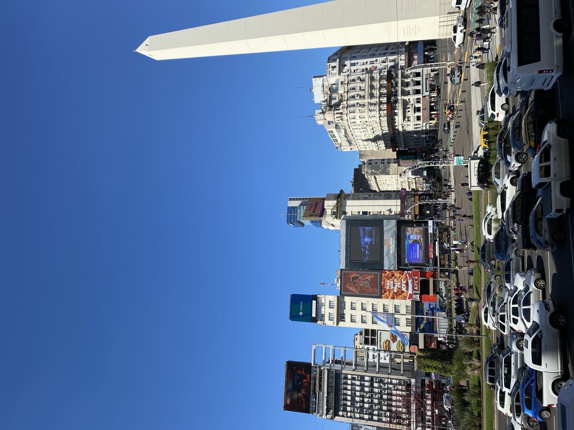 The image depicts the Obelisco de Buenos Aires, a national historic monument in the Plaza de la República, Buenos Aires, Argentina. The obelisk is in the upper left of the frame, contrasting with the clear blue sky. A line of buildings and billboards stretches along the middle of the frame. Below them are cars, buses, and pedestrian traffic in the foreground. The scene is bustling with activity.