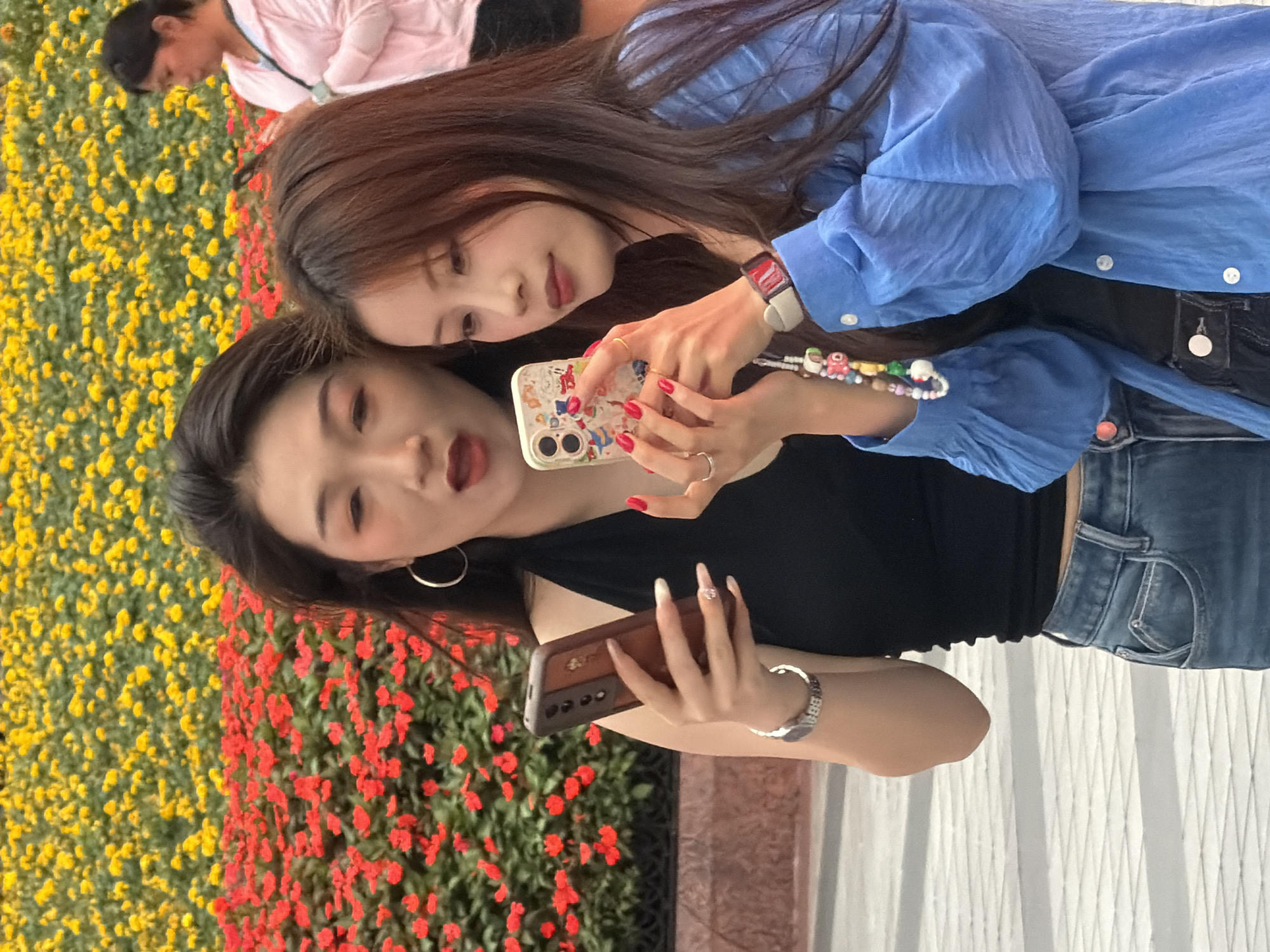 Two young East Asian women are in the foreground, looking at their smartphones. The woman on the left, wearing a black halter top and blue jeans, is holding a brown phone with her left hand. She has long dark hair and is wearing hoop earrings and a silver bracelet. The woman on the right, wearing a blue textured blouse and black top, is holding a white phone with a colorful case in her right hand. She has long brown hair and is wearing rings and a watch with a red band. Behind them, a flower bed is visible, with a layer of red flowers in the front and yellow flowers in the back. A third woman, partially visible, is in the background wearing a pink hooded jacket. The ground in front of the women is paved with a light-colored, patterned surface. The scene appears to be in an outdoor urban setting, potentially a park or public garden, in Yuzhong District, China.