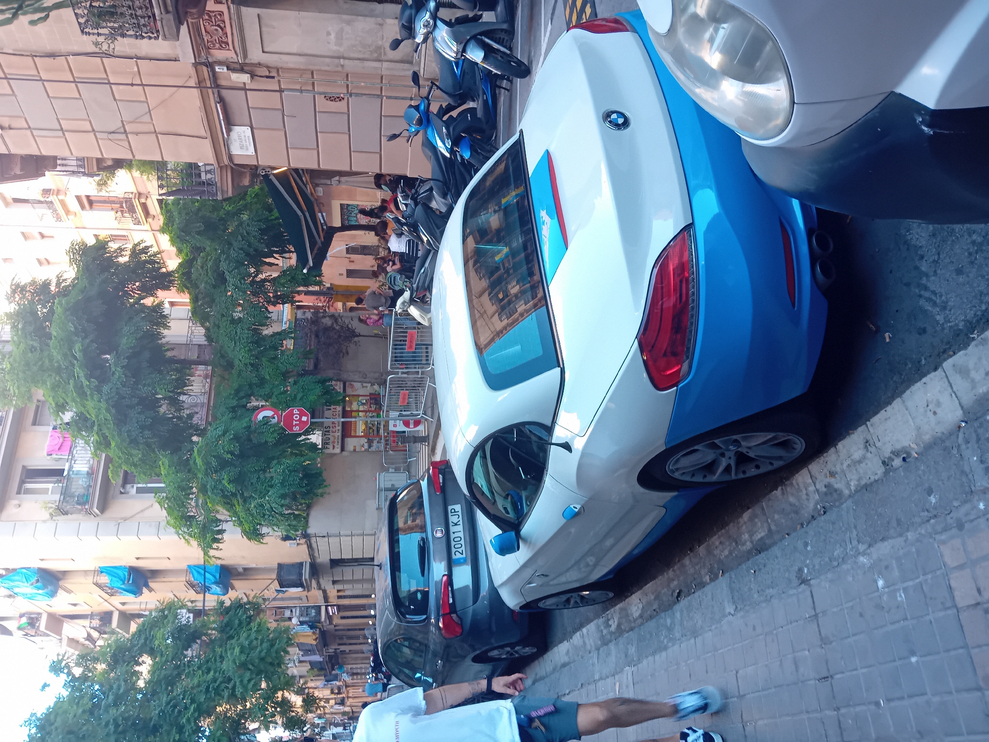 The image depicts a street scene in Barcelona, Spain. Parked cars line the sidewalk, including a white and blue BMW roadster and a grey Fiat. A group of people is gathered on the sidewalk near a storefront shaded by a green awning. Trees line the street, interspersed with buildings of various heights and architectural styles. A street sign, "PIZARRO," is visible on the corner of one of the buildings. Other signs include "STOP" and advertisements for "FRUTS Y ESPECIAS." Several motorcycles are parked behind the white car. A person is walking in the foreground, visible from the waist down. The license plate on the Fiat reads "2001 KJP."