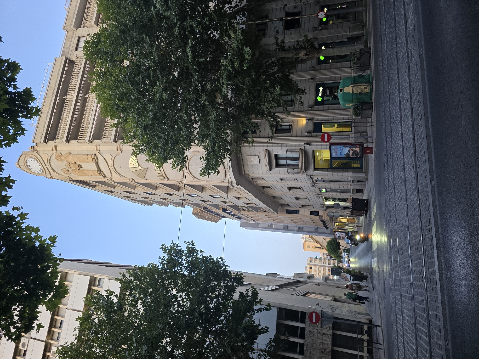 The image depicts a street scene in Jaén, Spain, under a clear sky at dusk. The foreground features tram tracks embedded in a cobblestone street. To the left, a modern building with large windows stands tall, partially obscured by leafy trees. Across the street, a light-colored, ornate building dominates the view, featuring balconies with railings, decorative reliefs, and a clock face near its peak.
Several trees with full, green foliage line the street, contributing to the urban landscape. In the middle ground, a motorbike with its headlights on is seen moving down the street, with a few pedestrians walking on the sidewalk. Buildings of varying architectural styles and heights are visible along the street, suggesting a city environment. A green recycling bin for glass is prominently placed on the sidewalk in front of a building with "Unicaja" signage. A red and white "no entry" sign is affixed to a pole on the left side of the street. Traffic lights and street lamps are also present. The overall atmosphere suggests a quiet urban evening.