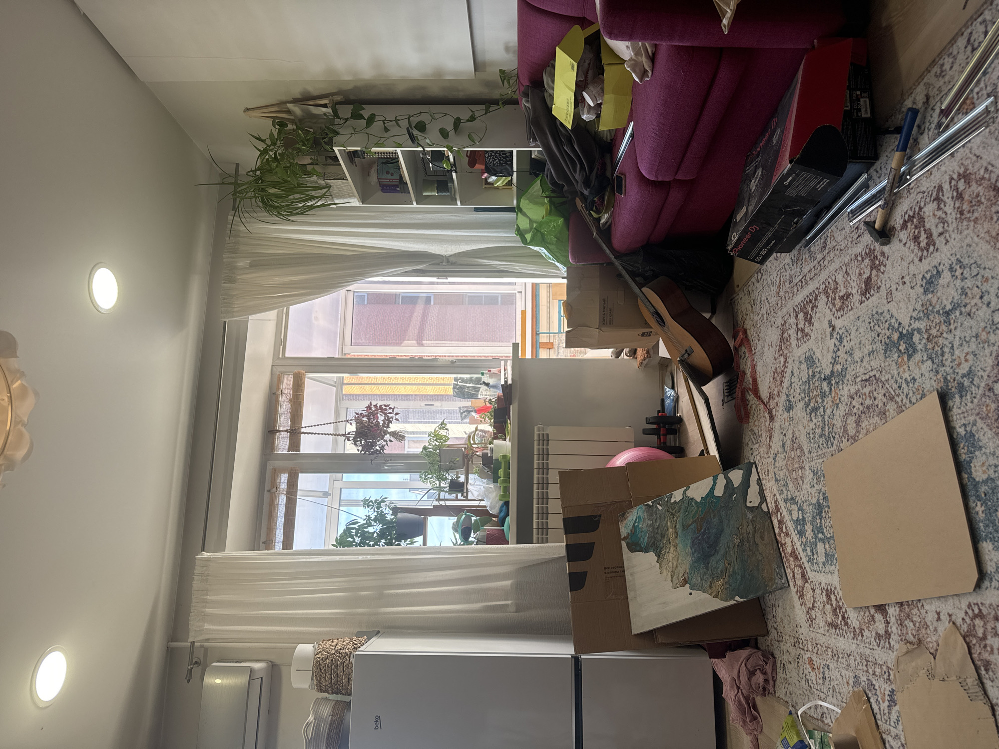 This is an interior shot, presumably of an apartment in Krasnoyarsk, Russia. The view is from the kitchen area towards a living space. The kitchen has a white refrigerator and a radiator near the window. The window is draped with white curtains and contains several potted plants. Beyond the window are buildings with red brick or painted facades, suggesting an urban residential area.
The living space appears cluttered. There's a magenta-colored sofa, a bookshelf, a guitar, various boxes, and equipment including a Pioneer DJ box. A rug with an intricate pattern covers the floor. Light fixtures are visible on the ceiling. Overall, the scene depicts a messy domestic environment.