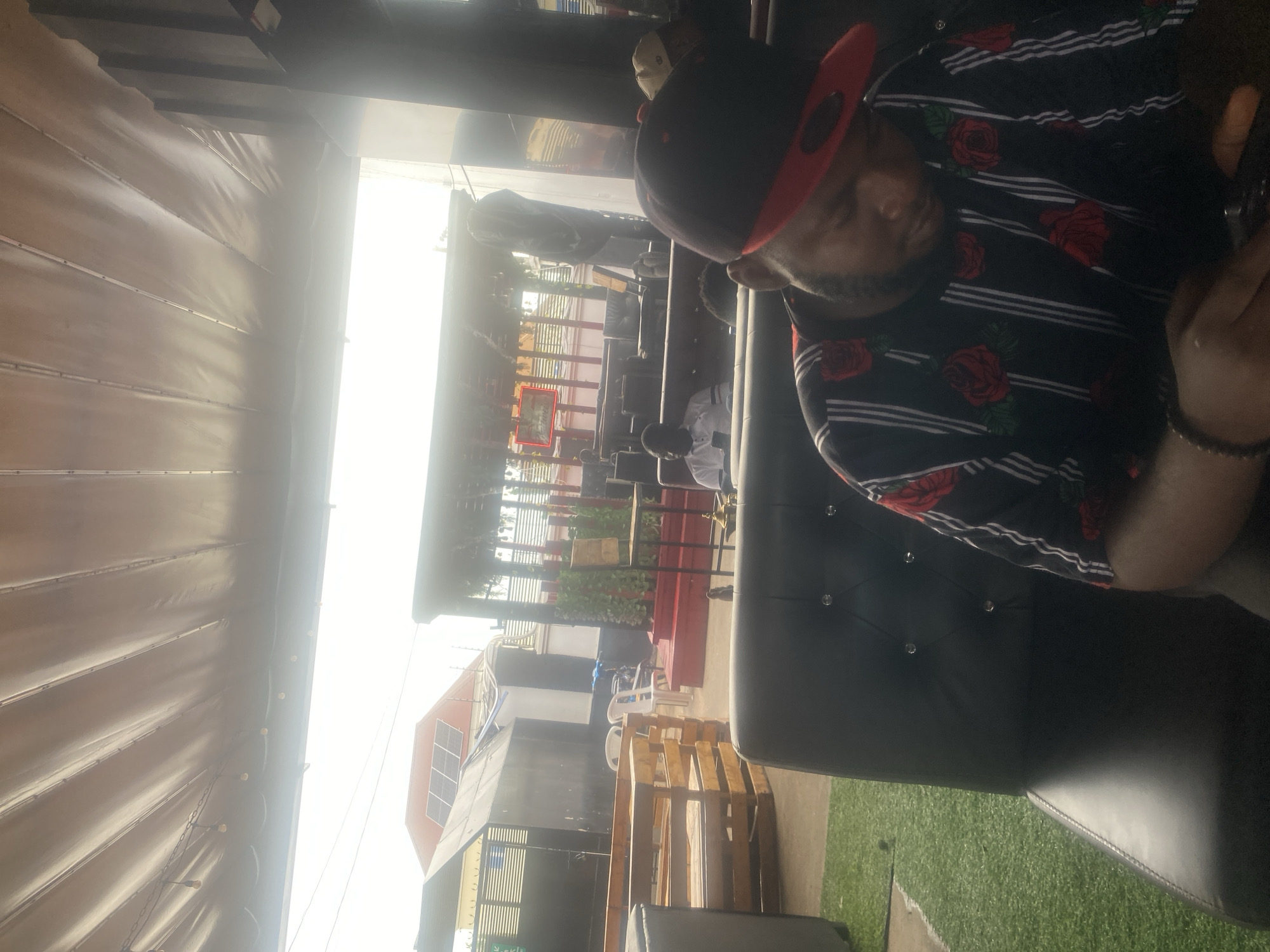 The image displays an outdoor or semi-outdoor commercial seating area located in Howayi, Nigeria, under bright daylight conditions. The upper portion of the frame is covered by a light-colored fabric canopy or awning from which string lights with individual bulbs are suspended.
In the foreground, a male individual is seated on a black, tufted, padded sofa. He is wearing a black long-sleeved shirt patterned with vertical white stripes and red roses. A black baseball cap with a red brim is worn backward on his head. He is observed looking downwards at a handheld electronic device partially visible in his hands. A dark beaded bracelet is present on his left wrist. The ground directly in front of the sofa is covered with artificial green turf.
In the mid-ground, to the left of the foreground subject, another male individual in a white shirt is seated on a similar black padded sofa, also engaged with a mobile phone. Further back on the right, the top of a third person's head, wearing a light-colored cap, is partially visible.
The environment beyond the immediate seating area includes a section with multiple wooden pallet constructions, potentially serving as benches or tables. Adjacent to these are two empty white plastic chairs. In the background, a dark fence with horizontal slats is visible, behind which stands a building with a red roof equipped with solar panels, and a smaller, dark, shed-like structure. Towards the right background, a red elevated platform or stage is present, adorned with green foliage. A dimly lit red sign and a small dark table holding what appears to be a hookah are also discernible. The sky in the background is significantly overexposed due to intense sunlight.