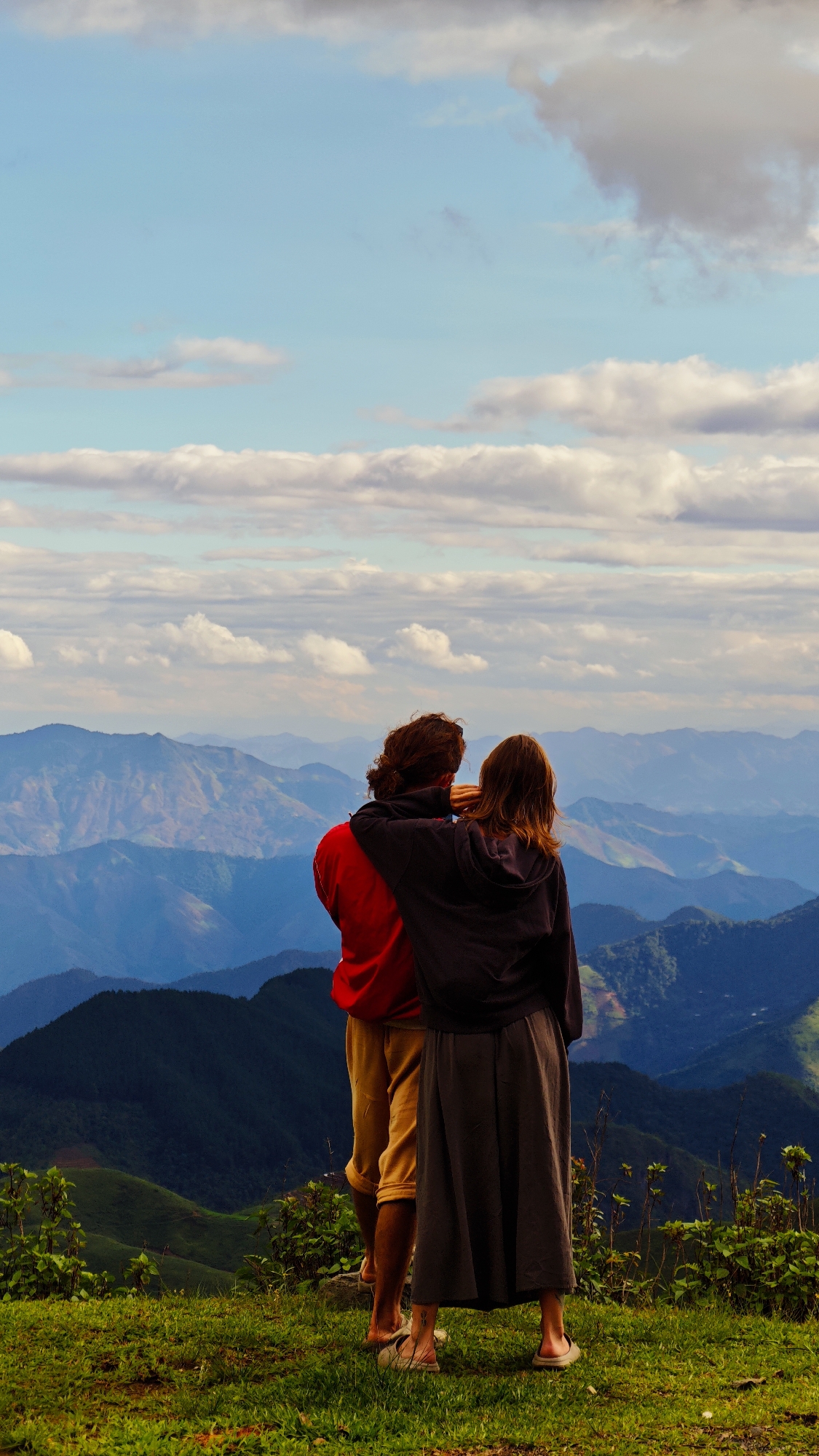 Two individuals are observed from behind, standing on a grassy hill overlooking a vast mountainous landscape under a partly cloudy sky in Xín Vàng, Vietnam. The person on the left, with long dark hair, wears a red long-sleeved top and mustard-yellow pants, with light-colored sandals visible. Their right arm is draped over the shoulders of the person on the right. The second individual, on the right, possesses brown, shoulder-length hair and wears a dark hooded sweatshirt over a long dark grey skirt, with light-colored flat shoes. Both individuals are positioned facing away from the camera, gazing out at the panoramic view. The foreground features a green grassy slope interspersed with small green plants. The midground and background consist of multiple layers of mountains, transitioning from darker green in closer proximity to lighter blue and hazy tones in the distance, indicating atmospheric perspective. The sky above is light blue and populated with scattered white and grey cumulus clouds of various sizes.