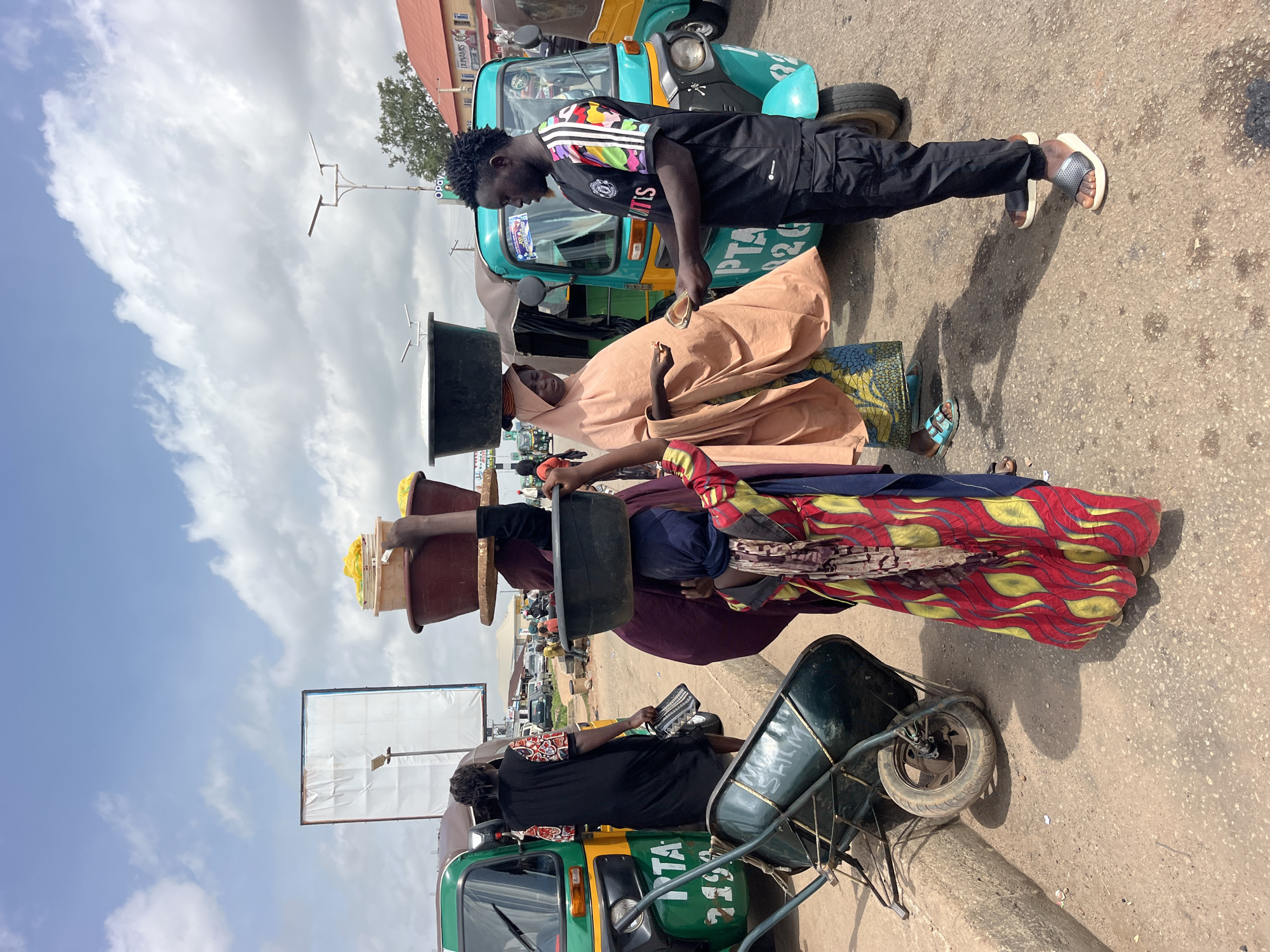 In the city of Unknown, Unknown, on a paved road under a cloudy sky, several people are engaged in commerce. A young man in a black shirt with colorful sleeves and black pants stands beside a turquoise auto-rickshaw, holding out money to a woman. This woman, wearing a peach-colored hijab and a patterned skirt, balances a large black basin on her head and appears to be receiving the money, holding out a small item in her hand. Another woman in a burgundy robe and a blue headscarf, also carrying a large basin on her head, stands behind the first woman, her body turned away from the viewer. To the left, a woman in a black dress with patterned sleeves walks away from the camera, carrying a patterned bag. A green wheelbarrow rests on its side near the auto-rickshaw. In the background, more auto-rickshaws and buildings are visible.