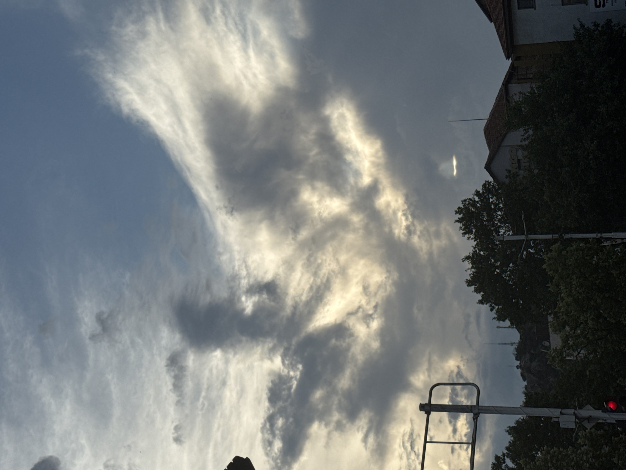 The image depicts a cloudy sky above a city, likely Plovdiv, Bulgaria, given the provided context. In the foreground, a traffic light is visible with a red signal illuminated. To the right of the traffic light, a metal pole with some equipment attached is present. The sky is filled with dramatic clouds, some dark and ominous, others illuminated by sunlight breaking through, creating a sense of depth and movement. In the mid-ground, the tops of trees are visible, and behind them, buildings with reddish-brown tiled roofs can be seen. A rocky hill or mountain with several communication towers is also discernible in the distance, suggesting a mountainous terrain within or near the city. The overall atmosphere is overcast, with a mix of dark and bright areas in the sky.