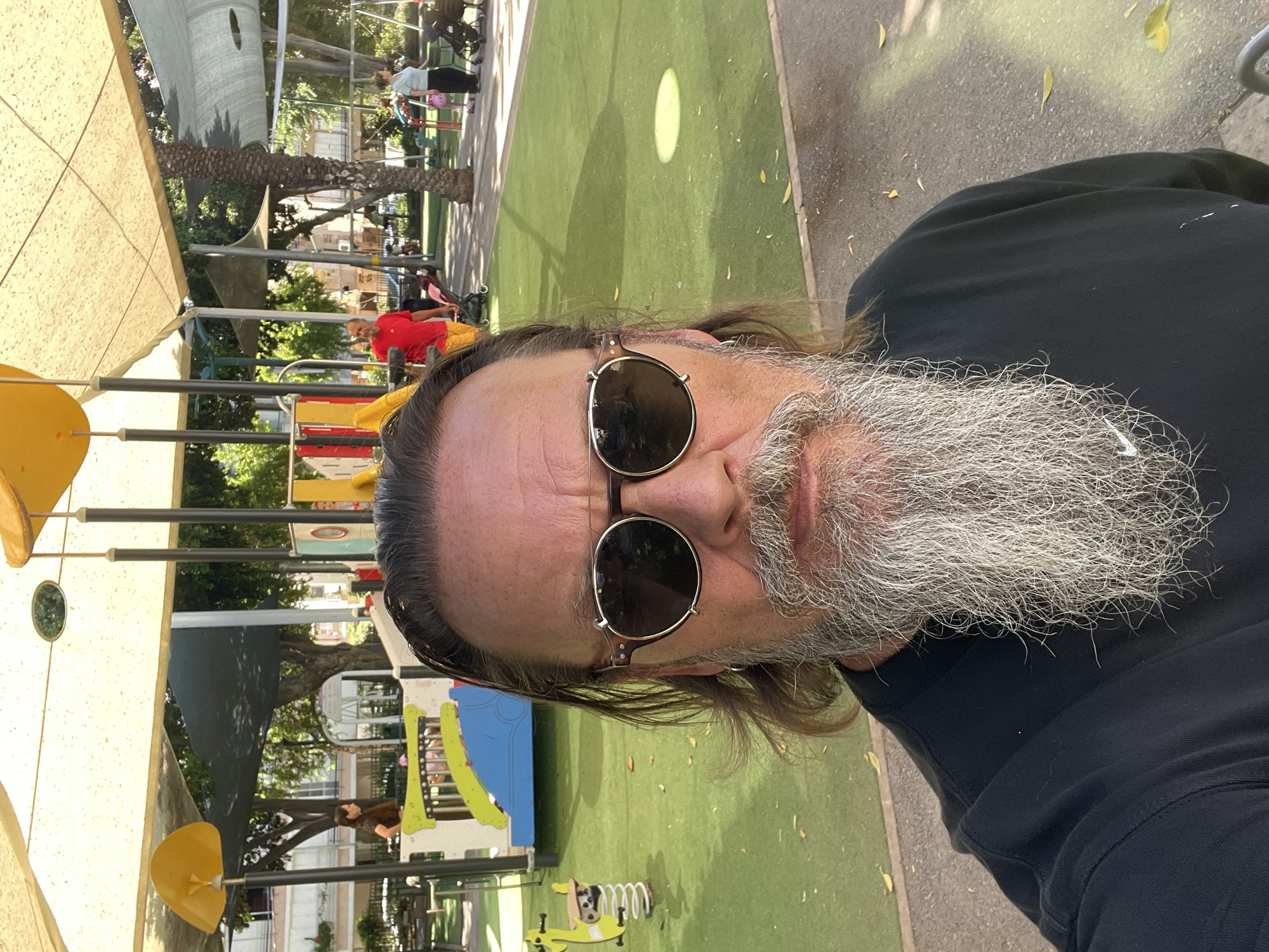 A man with long gray hair, a full gray beard, and sunglasses is taking a selfie in an outdoor playground. He is wearing a black sweatshirt. In the background, there is playground equipment, including slides and swings, and several other people, including adults and children, are visible. The ground is covered with green artificial turf and some paved areas. The playground appears to be located in a city, with buildings visible in the distance. The lighting suggests it is daytime and sunny. The overall impression is a casual, public space in an urban setting, likely a park or playground. The image was taken in Netanya, Israel.